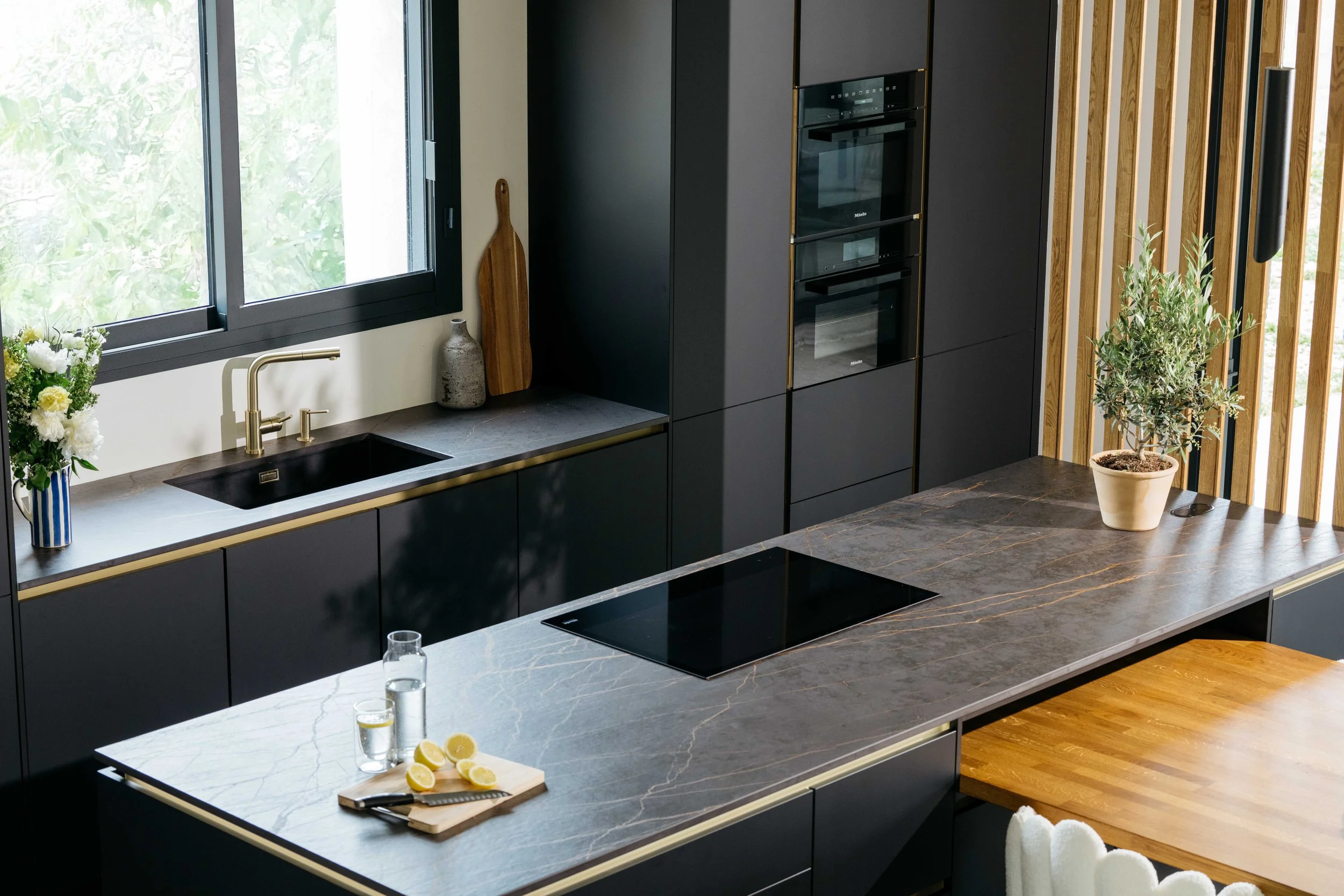 Moderne kök med mörka skåp, en köksö med marmorskiva, fönster med utsikt och några växter på bordet.