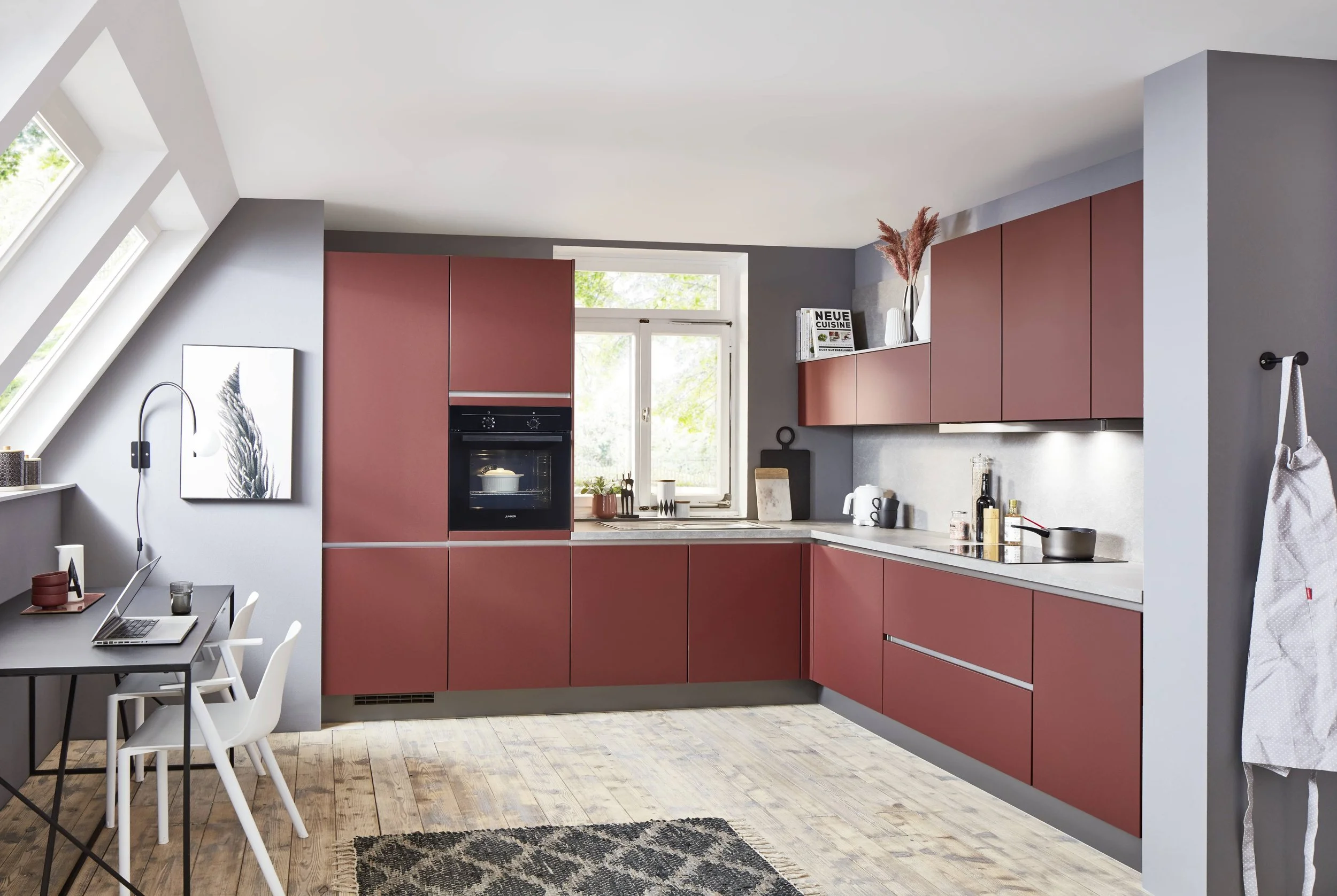 Kök med röda skåp, svart diskbänk, köksutrustning, bord med laptop, stolar, fönster och växter