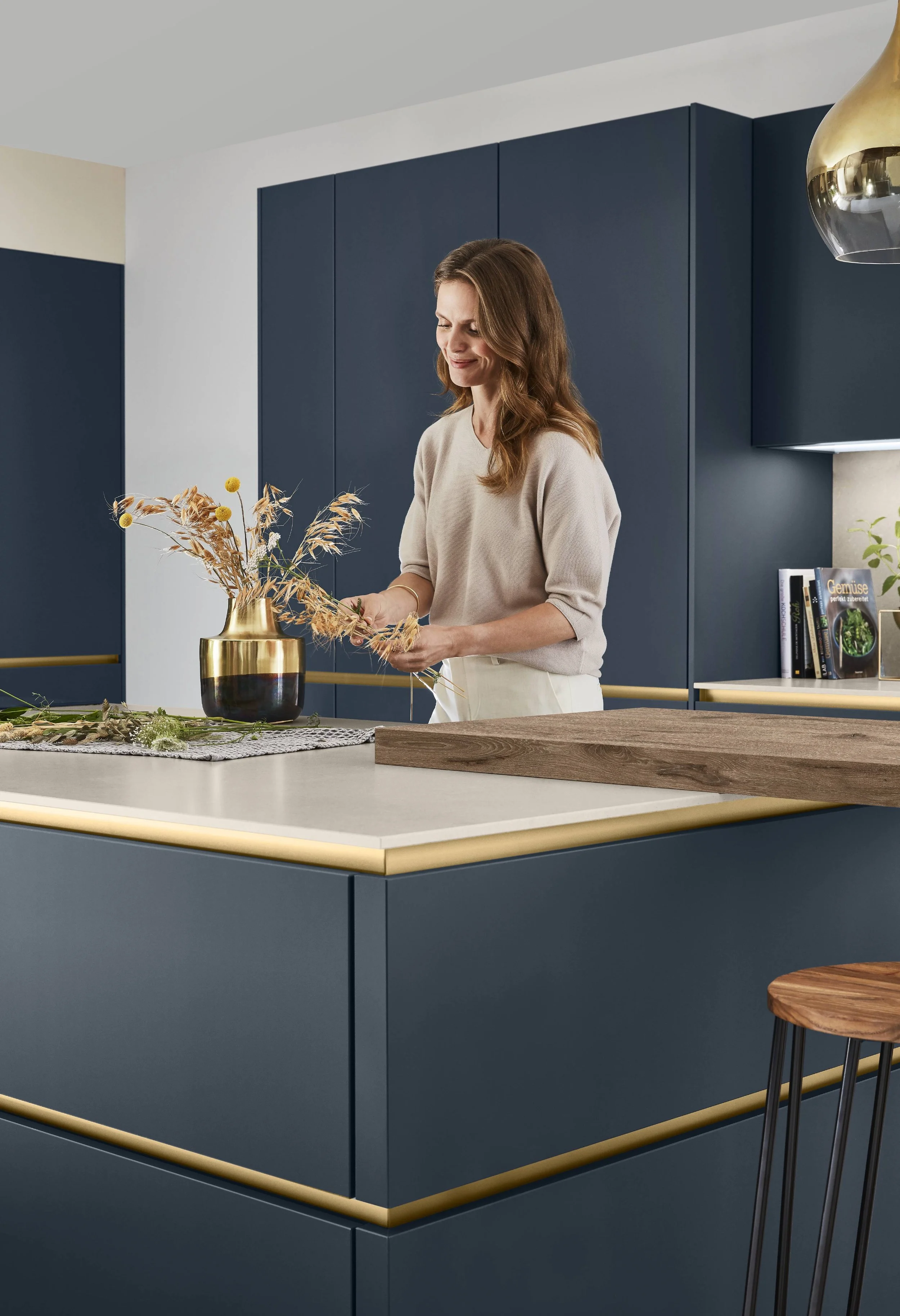 En kvinna står i ett modernt kök och arrangerar en bukett med torkade blommor i en guldfärgad och svart vas.
