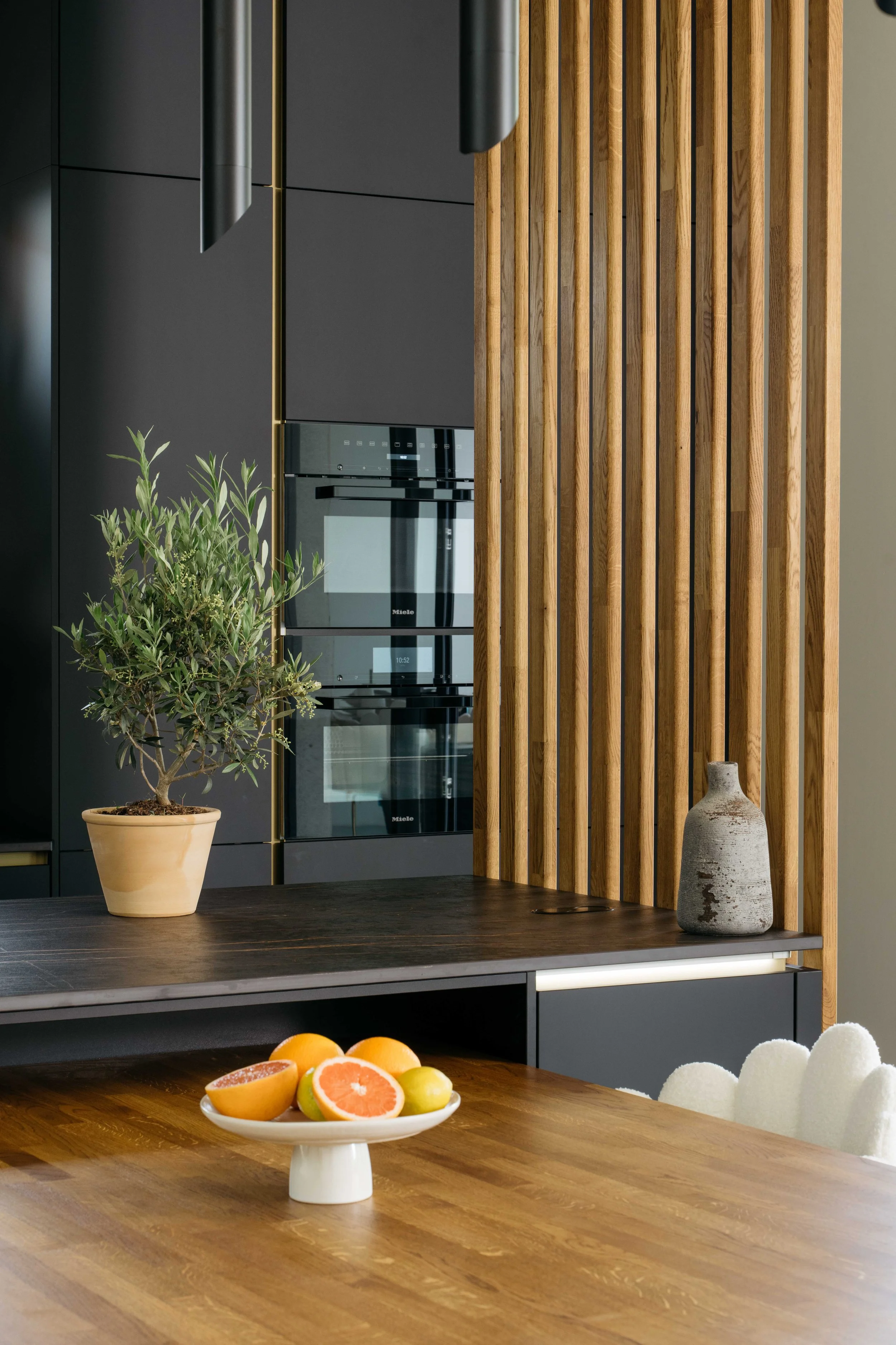Kök med svarta skåp, träluckor, ett bord med fruktskål, ett grönt växt i kruka och en keramisk vas.