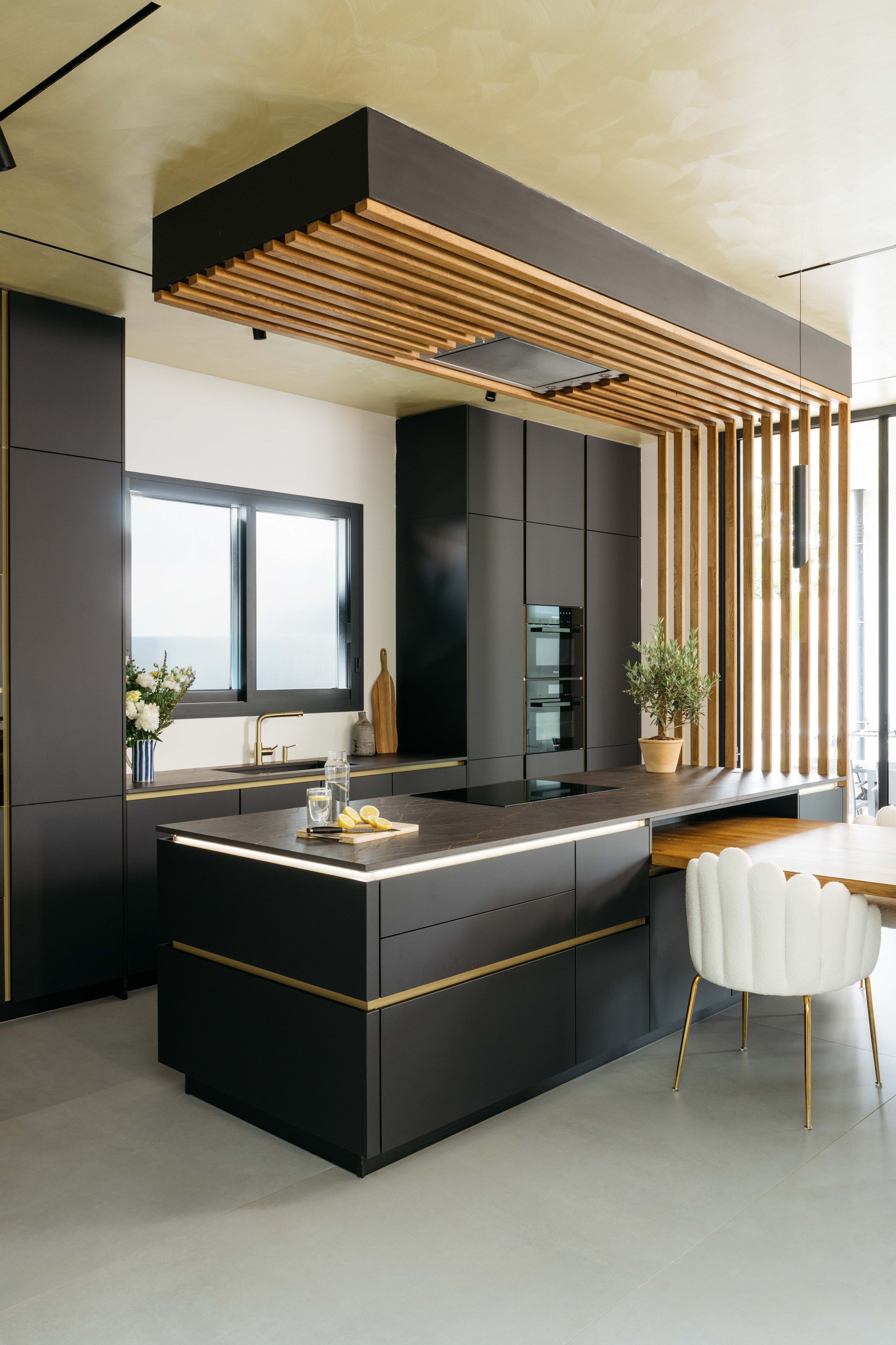 Modern svart kök med trämöbler och valv, köksö med ljuslist runt kanten, fönster och vita stolar.