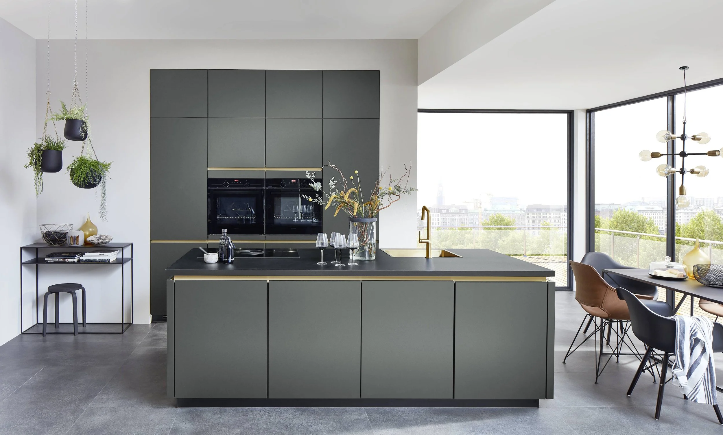 Modern kök med grå skåp och köksö, utsikt mot stad med stora fönster, svarta stolar och ett matbord, gröna växter och dekorationsföremål.