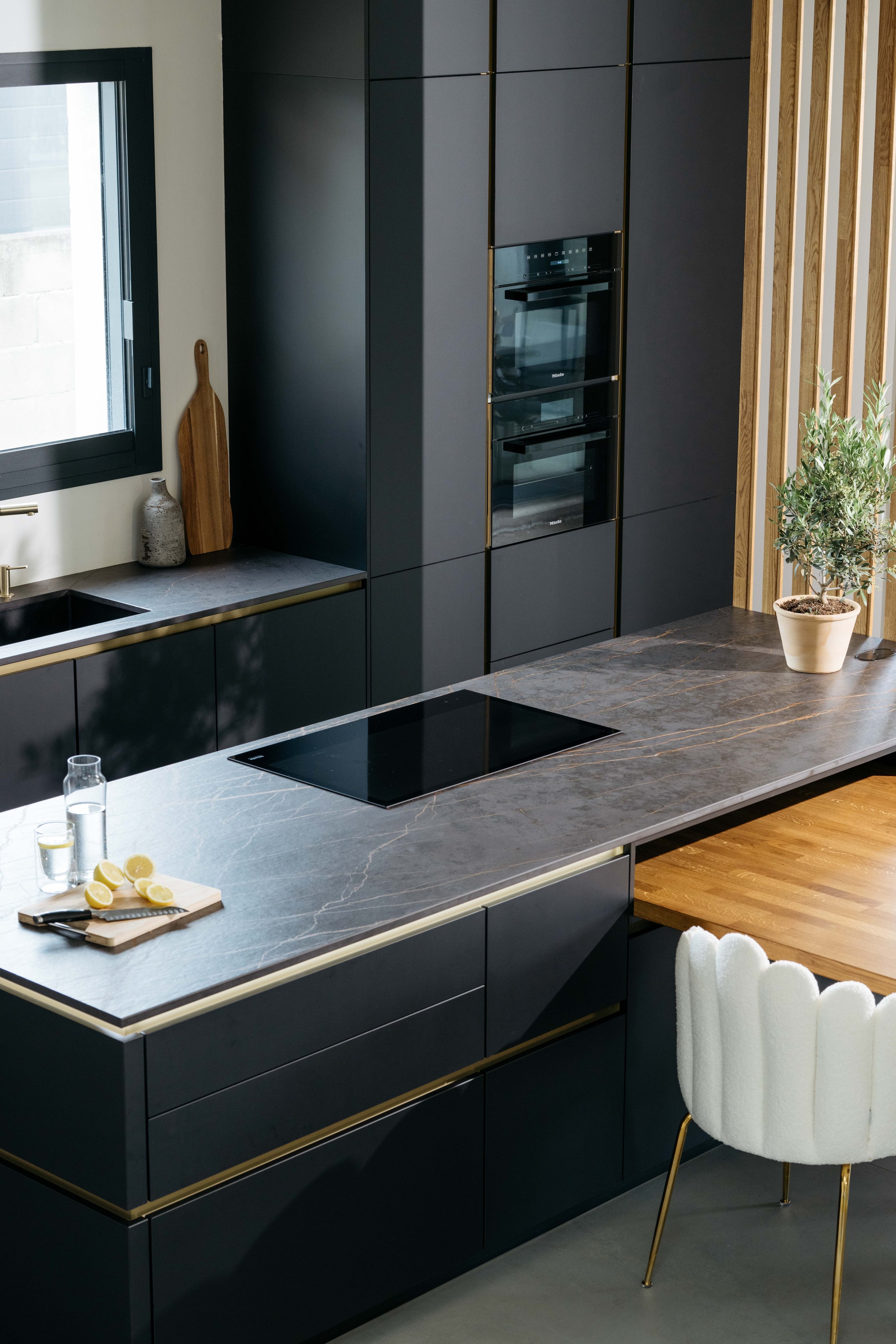 Modern köksinredning med svarta skåp och en kokplatta på en mörk marmorskiva. Det finns en fönster till vänster, några köksredskap och en växt på köksbänken.