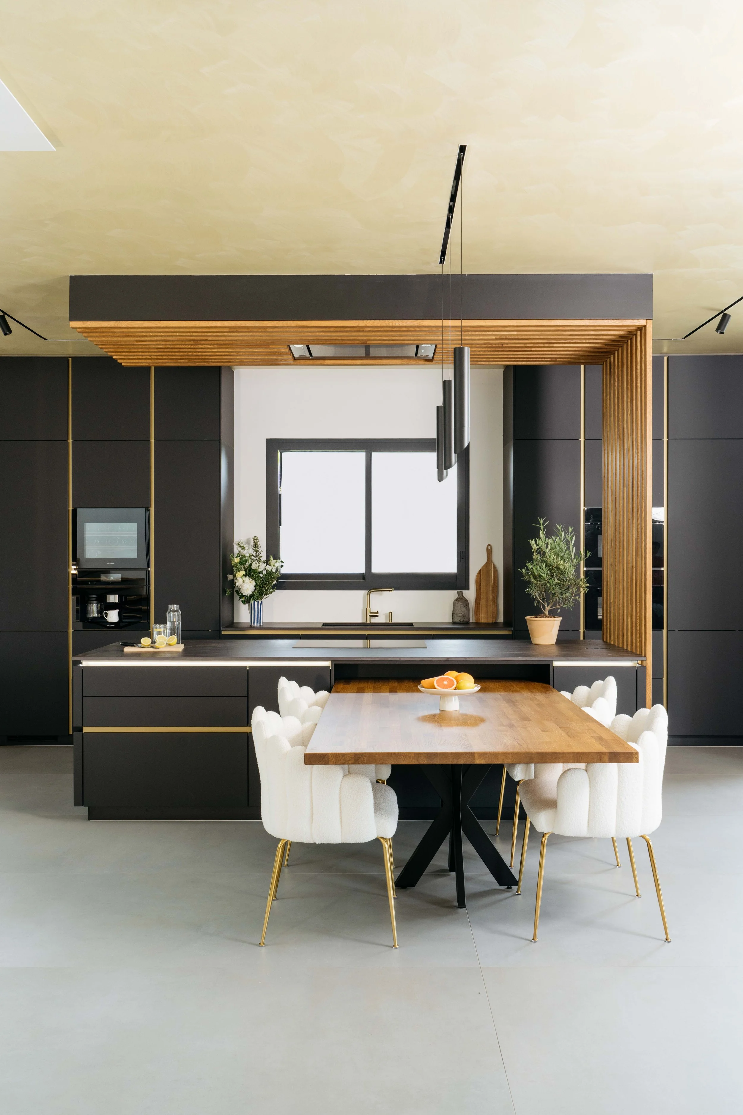 Modern kök med svart skåp, trädetaljer, ett fönster ovanför vasken, ett matsalsbord med vita stolar, och dekorationer som blommor och frukter.