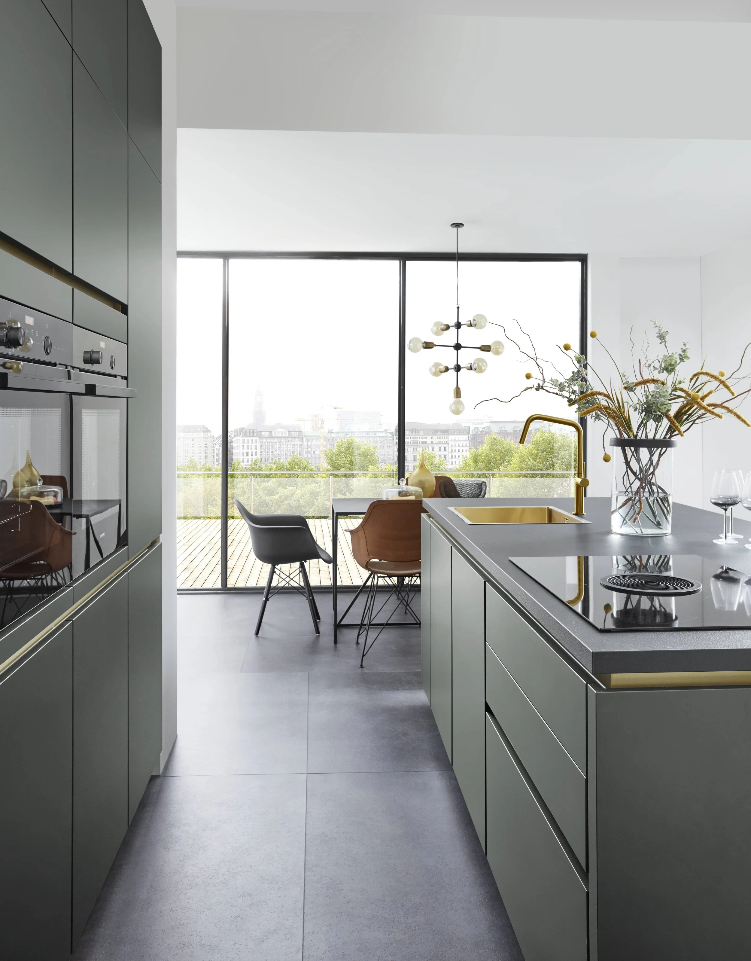En modern köksö med en guldfärgad diskho och kran, utsikt över en stad från ett stort fönster, dekorerad med en stor vas med blommor. Bänkar, stolar och ett bord är placerade nära fönstret.