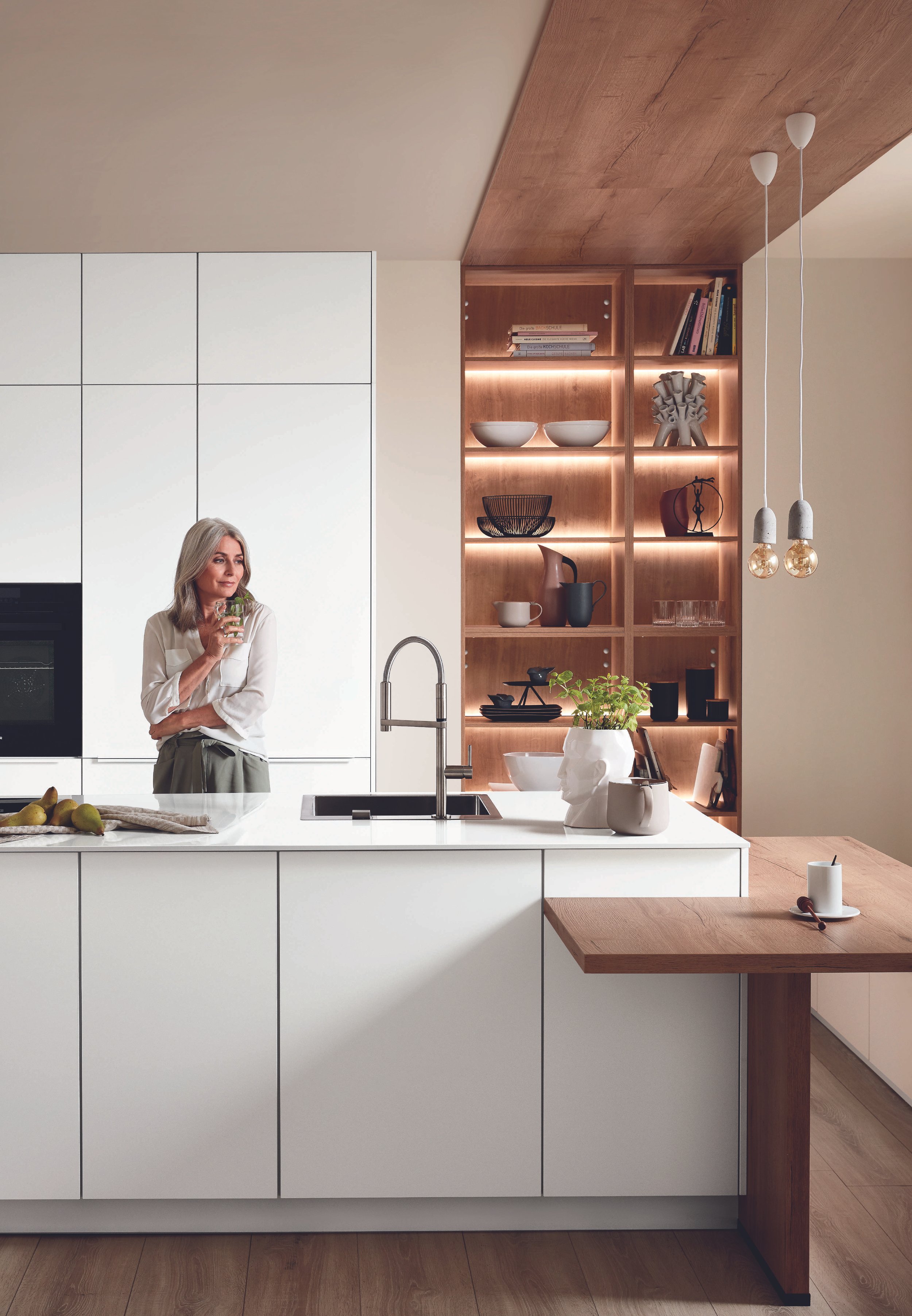 En kvinna står i ett modernt kök med vita skåp och en köksbänk. Det finns en växt i en vit skulptural kruka på köksbänken. I bakgrunden finns ett trävitrinskåp med böcker och dekorativa föremål.