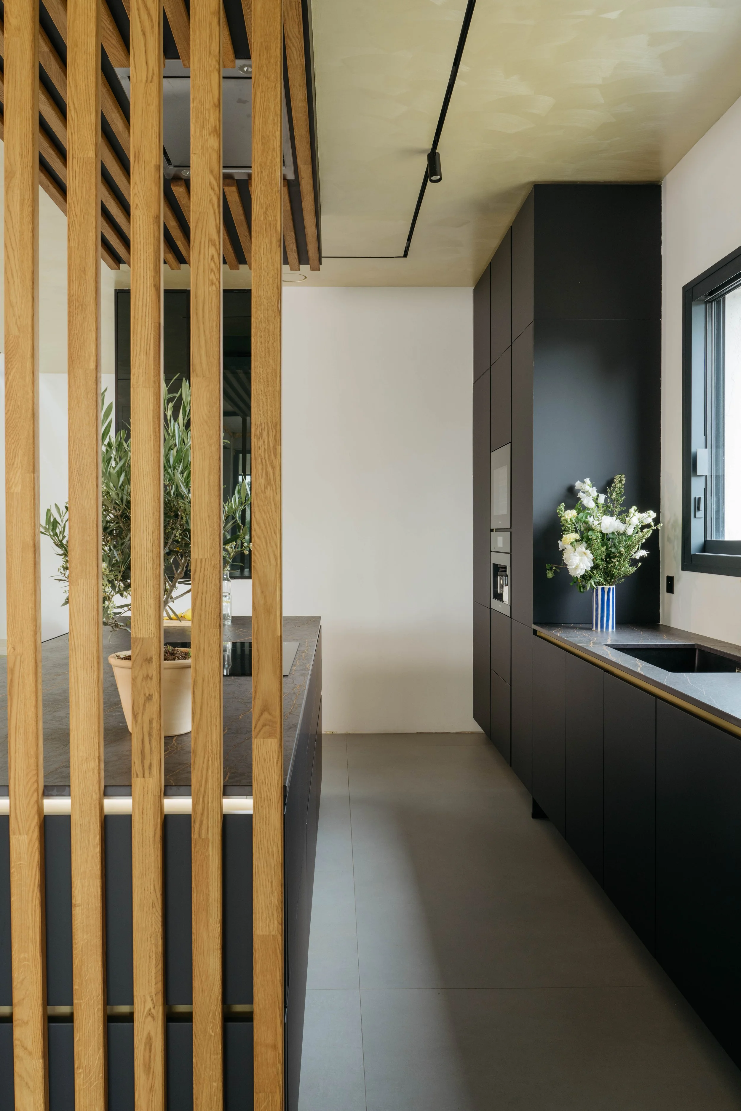 Modernt kök med svarta skåp, en begränsad planlösning, ett fönster, blombukett på bänken, och en träskärm som skiljer av köket från rummet.