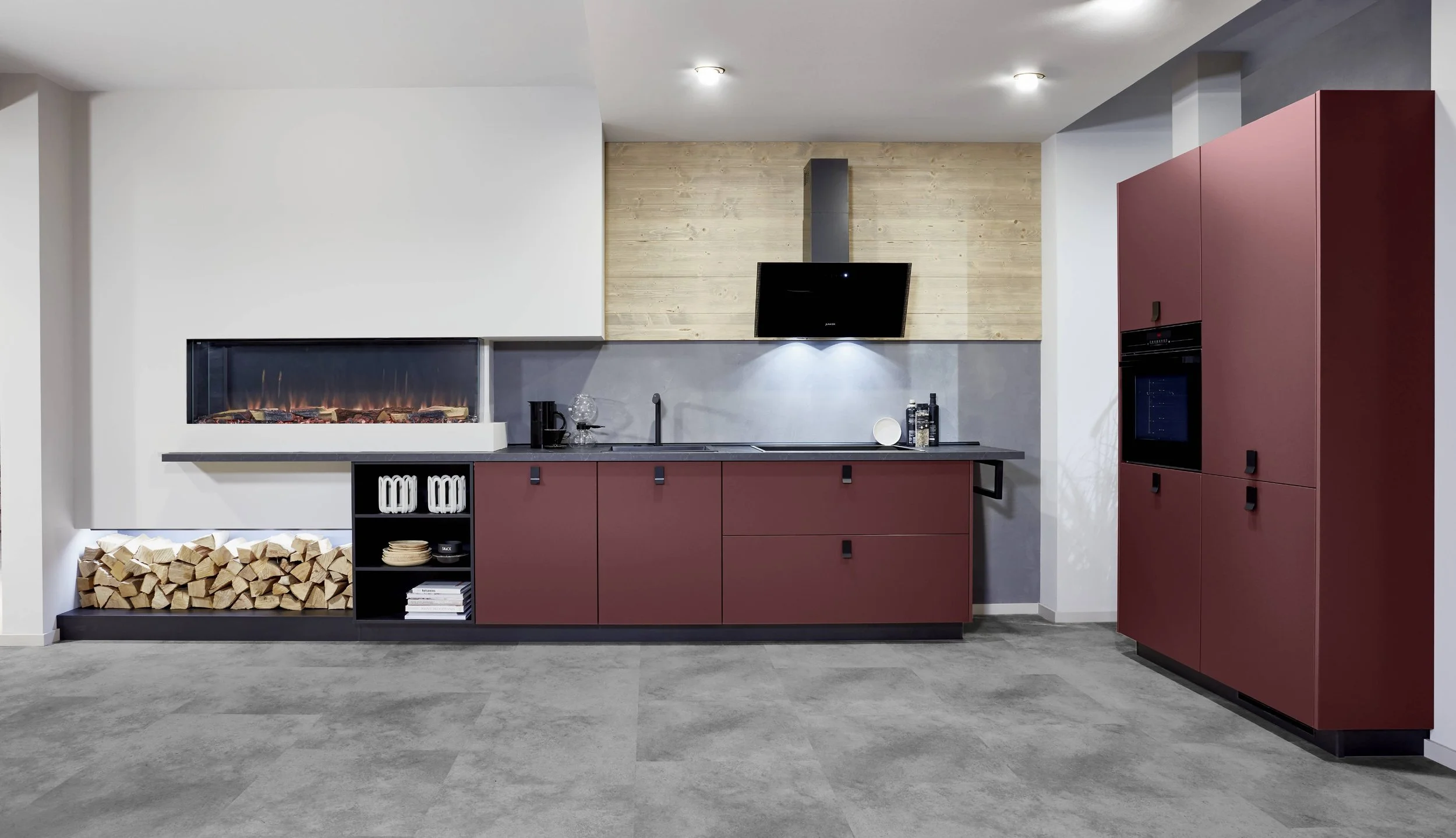 Modernt kök med röda skåp, öppen spis, vedförvaring, svart bänkskiva och inbyggd ugn, träpanel på väggen bakom spisen, och en platt-tv ovanför.
