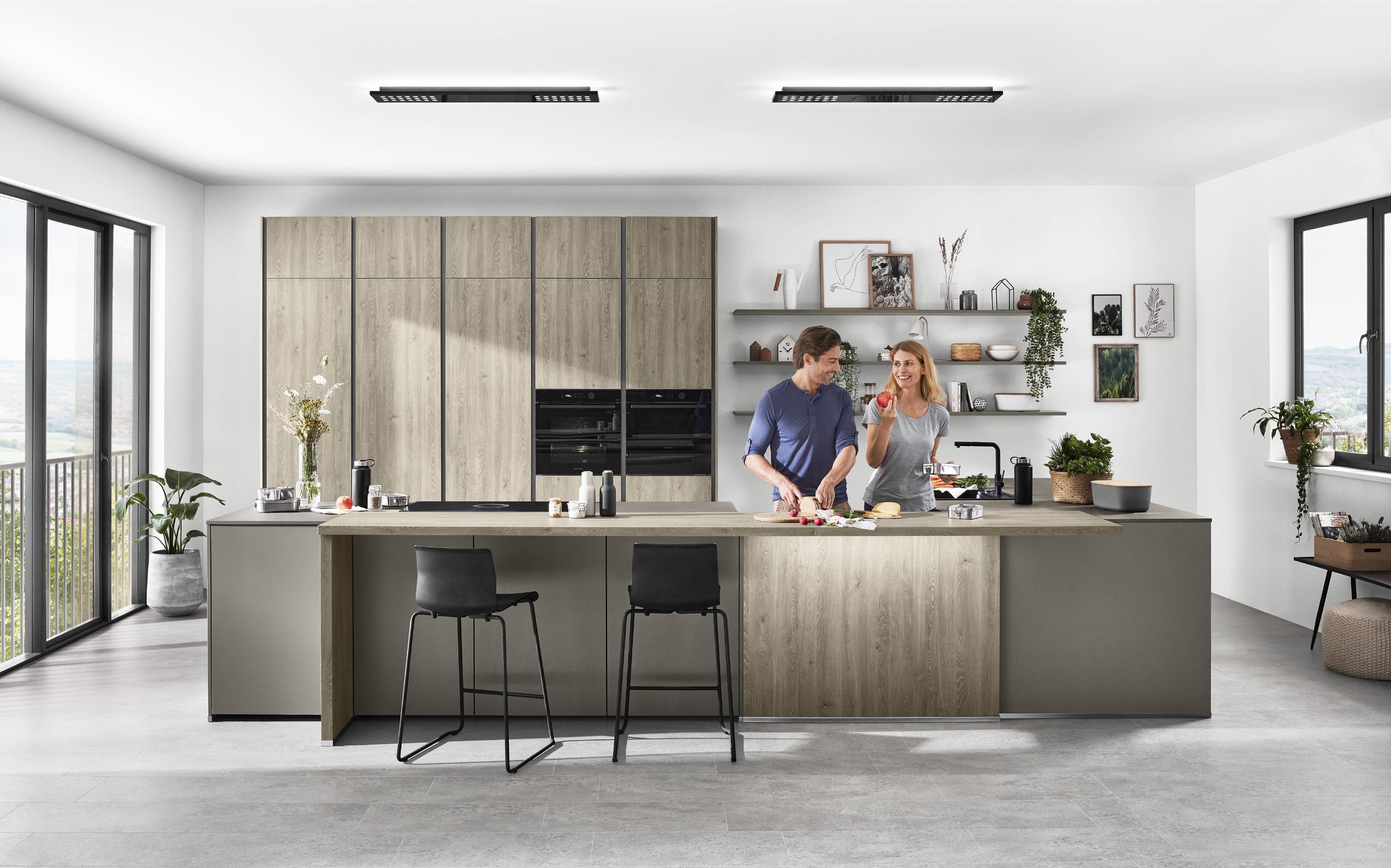 Ett modernt kök med två personer som lagar mat och äter äpplen, stora fönster och avskalad inredning.