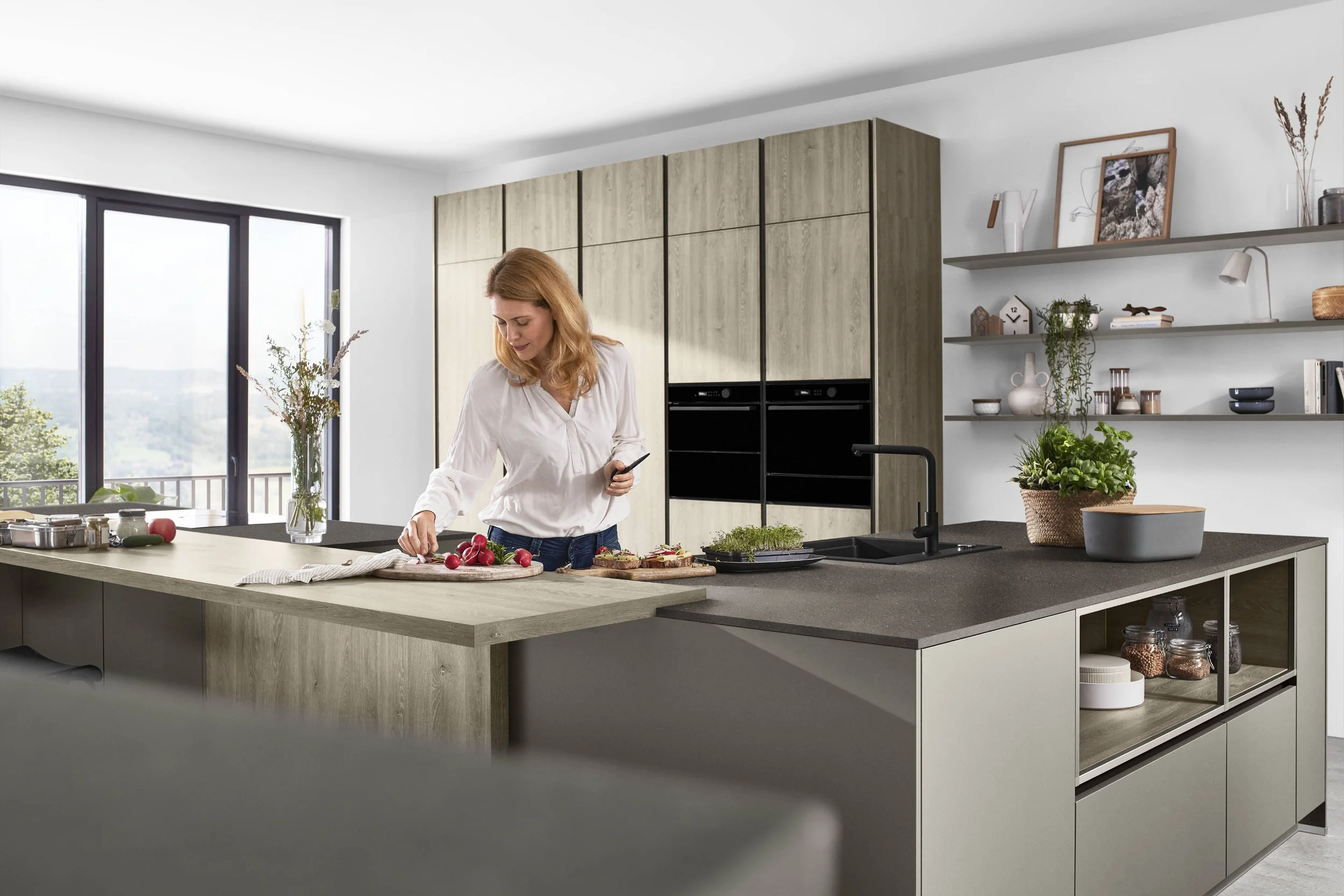 en kvinna som skär grönsaker i ett kök med moderna skåp och hyllor, stort fönster med utsikt
