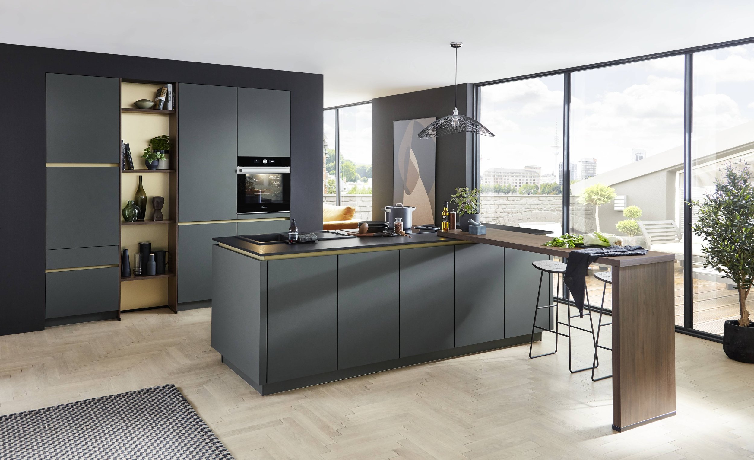 Modern köksmiljö med mörkgröna skåp, ett köksö, en stor glasvägg som visar en uteplats med balkongmöbler och gröna växter.