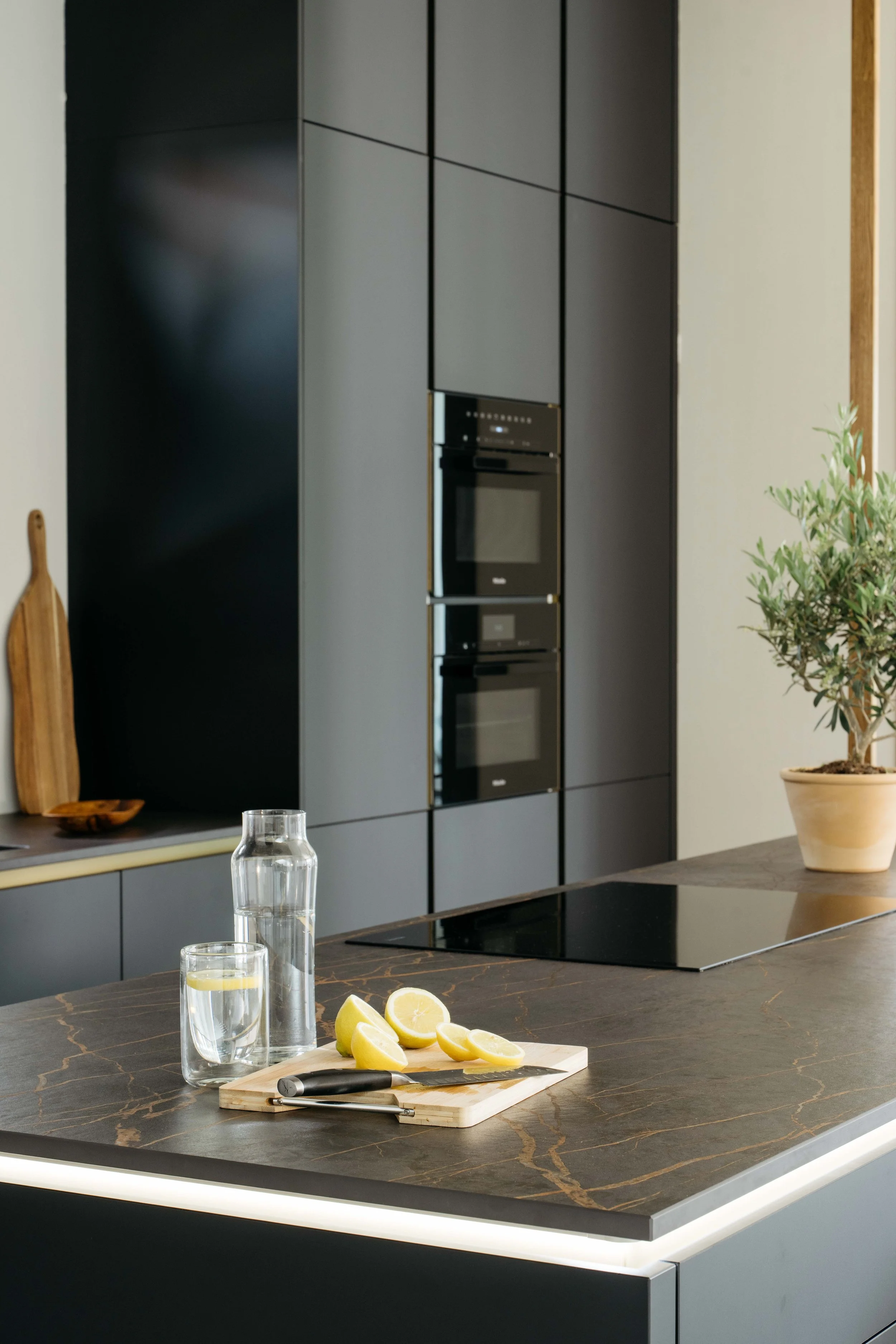 Köksö med lemon wedges, ett glas vatten, en vattenkaraff, en kniv och ett skärbräda, modern köksinredning med svarta och grå skåp och inbyggd ugn, grönt växt i kruka.
