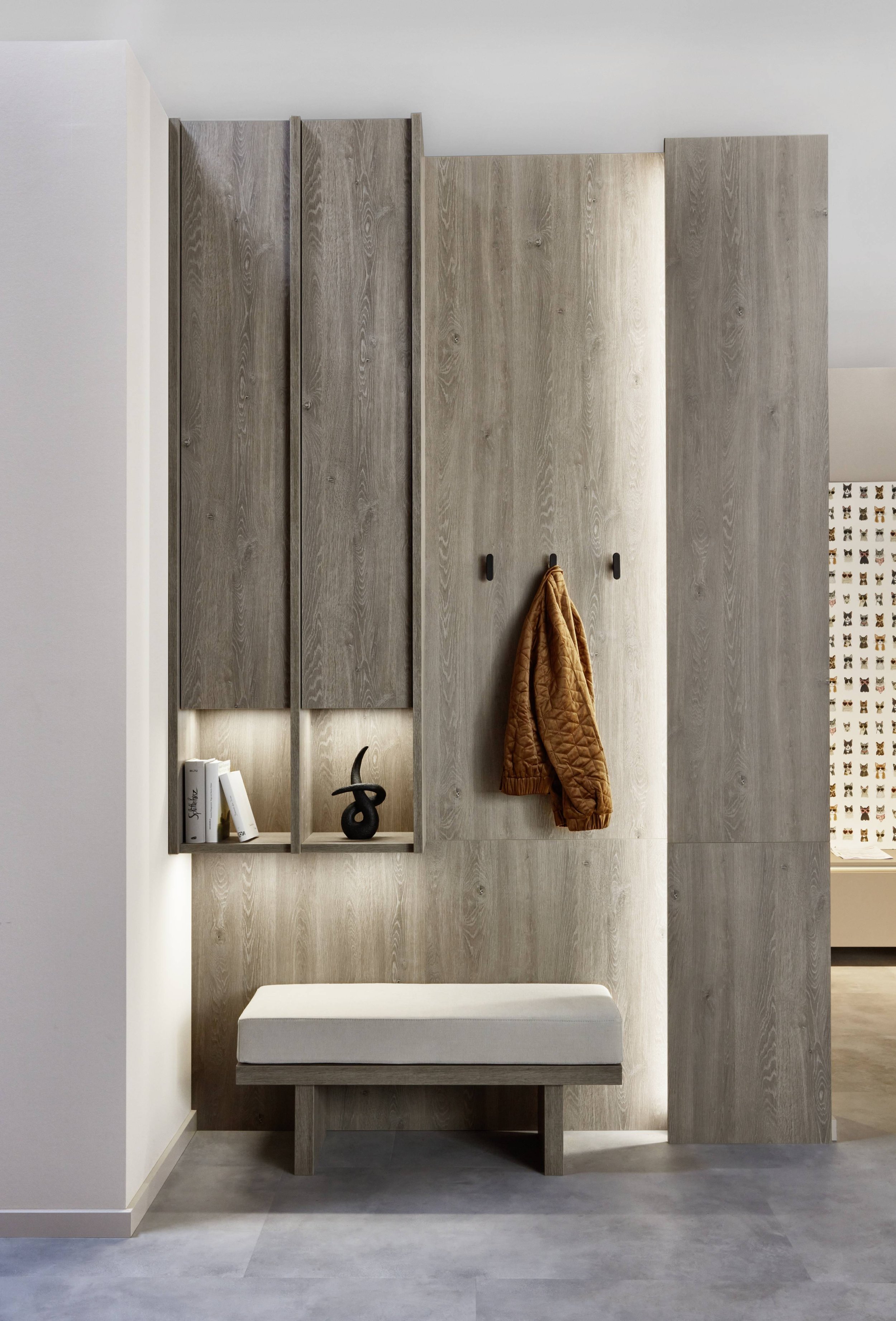 En modern hall med väggskåp i ljus och mörk trä. En bänk med sittdyna under ett vägghängt klädhängare med en brun jacka. På hyllan finns några böcker och en svart dekorativ skulptur.