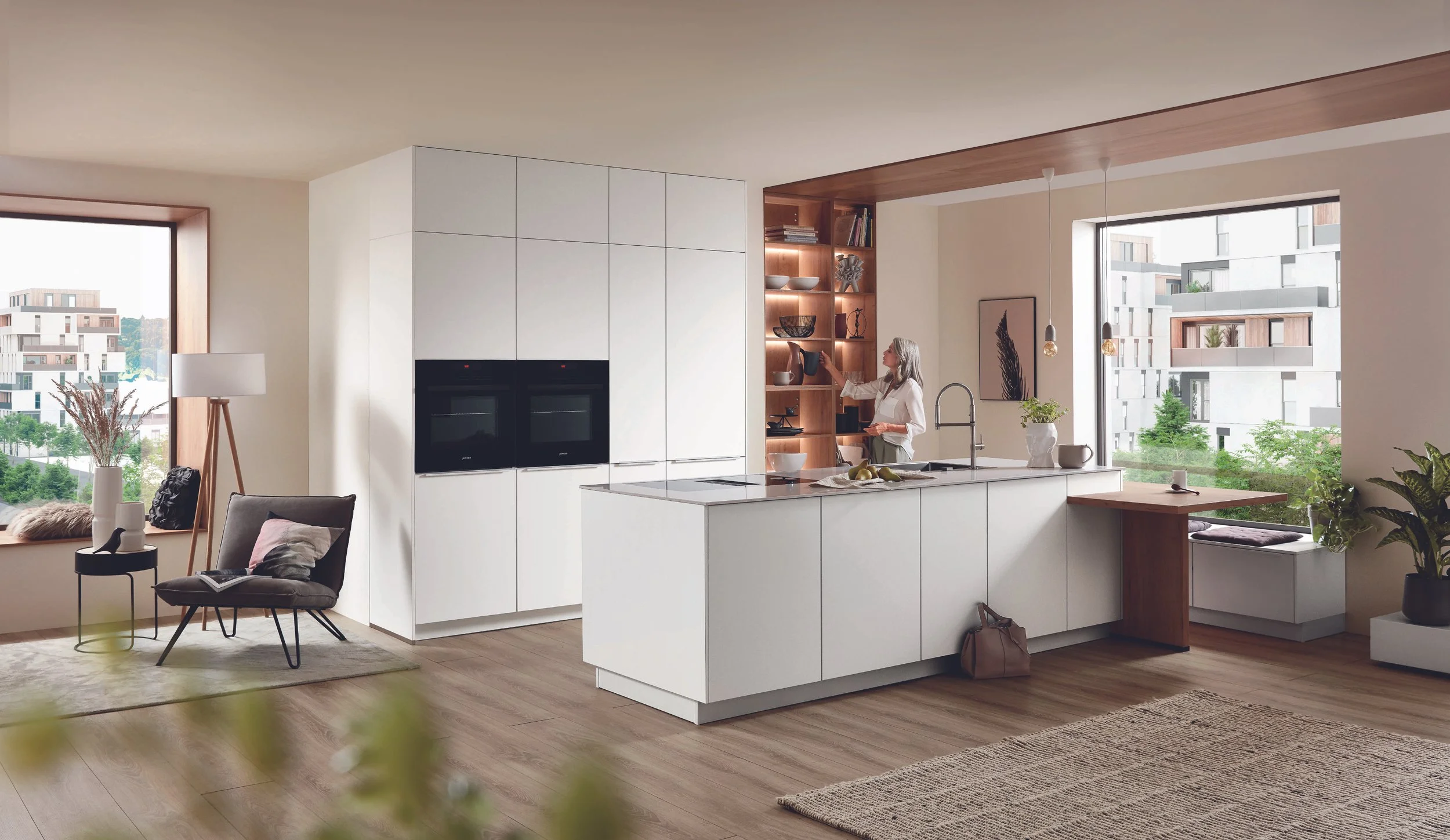 Modern kök med stor köksö, inbjudande sittplats vid fönster, och en kvinna som tittar på hyllor. Stora fönster med utsikt över moderna byggnader och grönska.