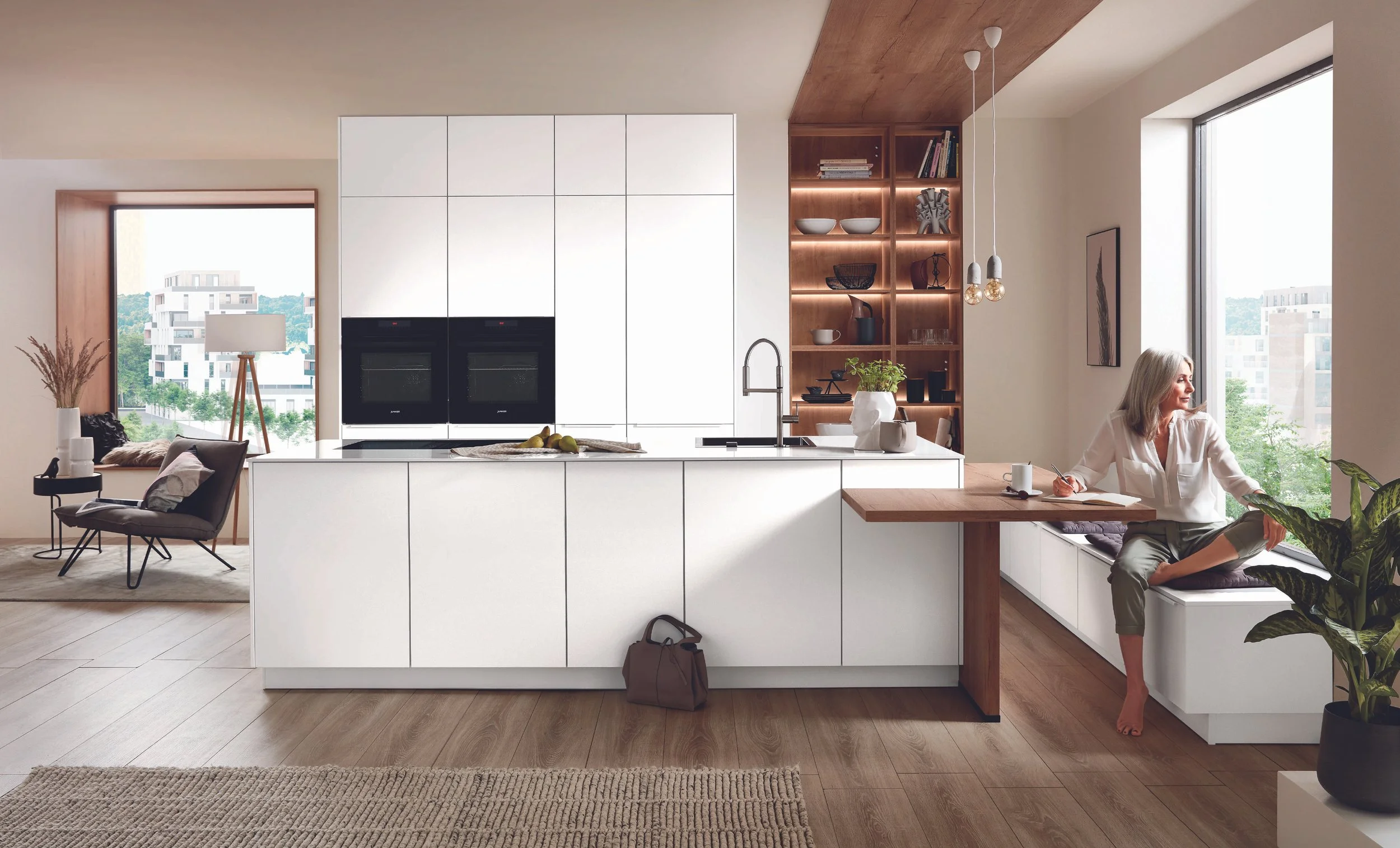 Modern köks- och vardagsrum med vit köksö, öppen hylla med prydnadsföremål, kvinna som sitter vid fönsterbänken och läser