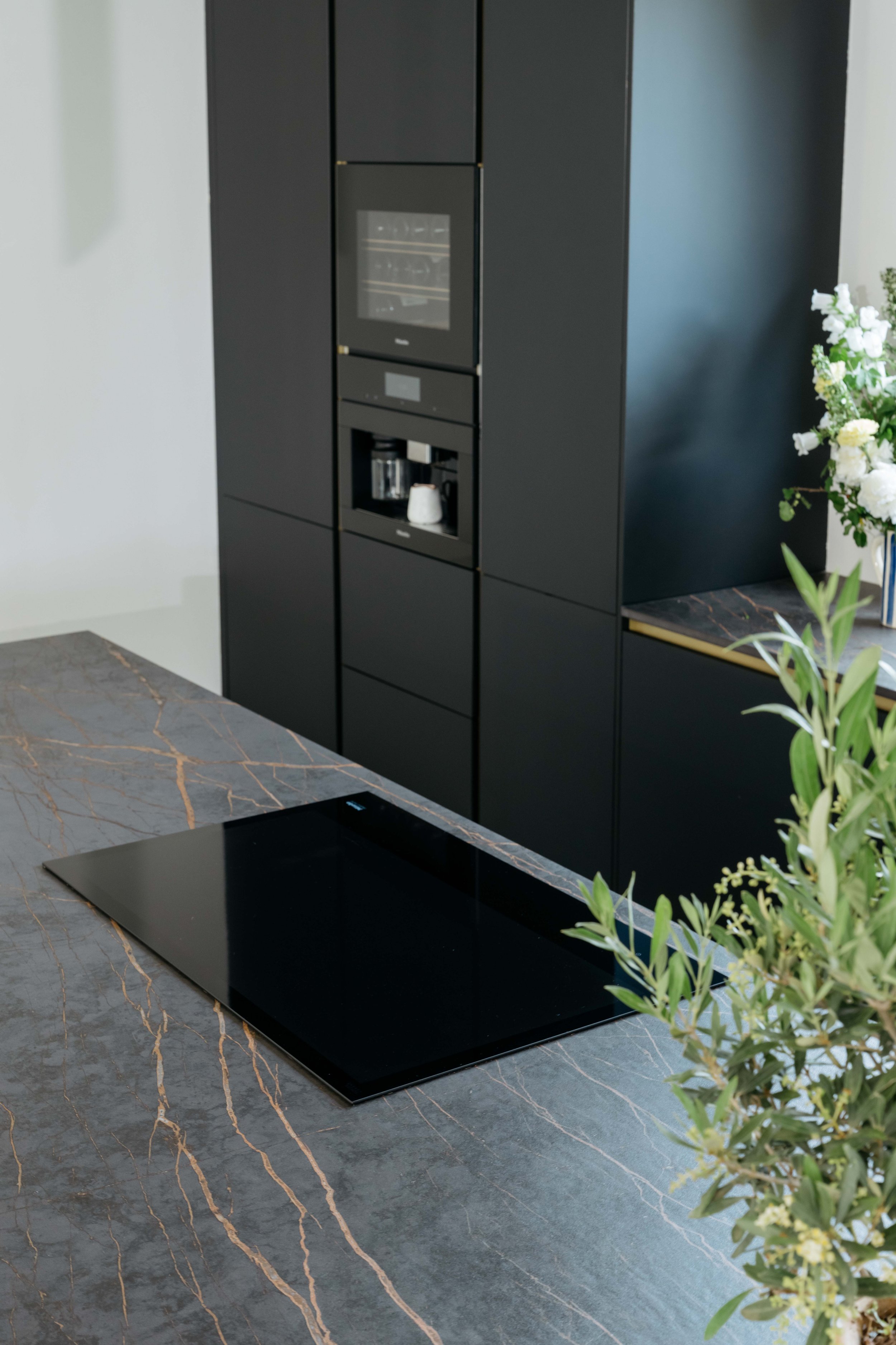 En modern köksö med en svart glaskeramikhäll och en grå marmorskiva. Bakom finns ett svart skåp med inbyggda ugnar och en kaffemaskin.