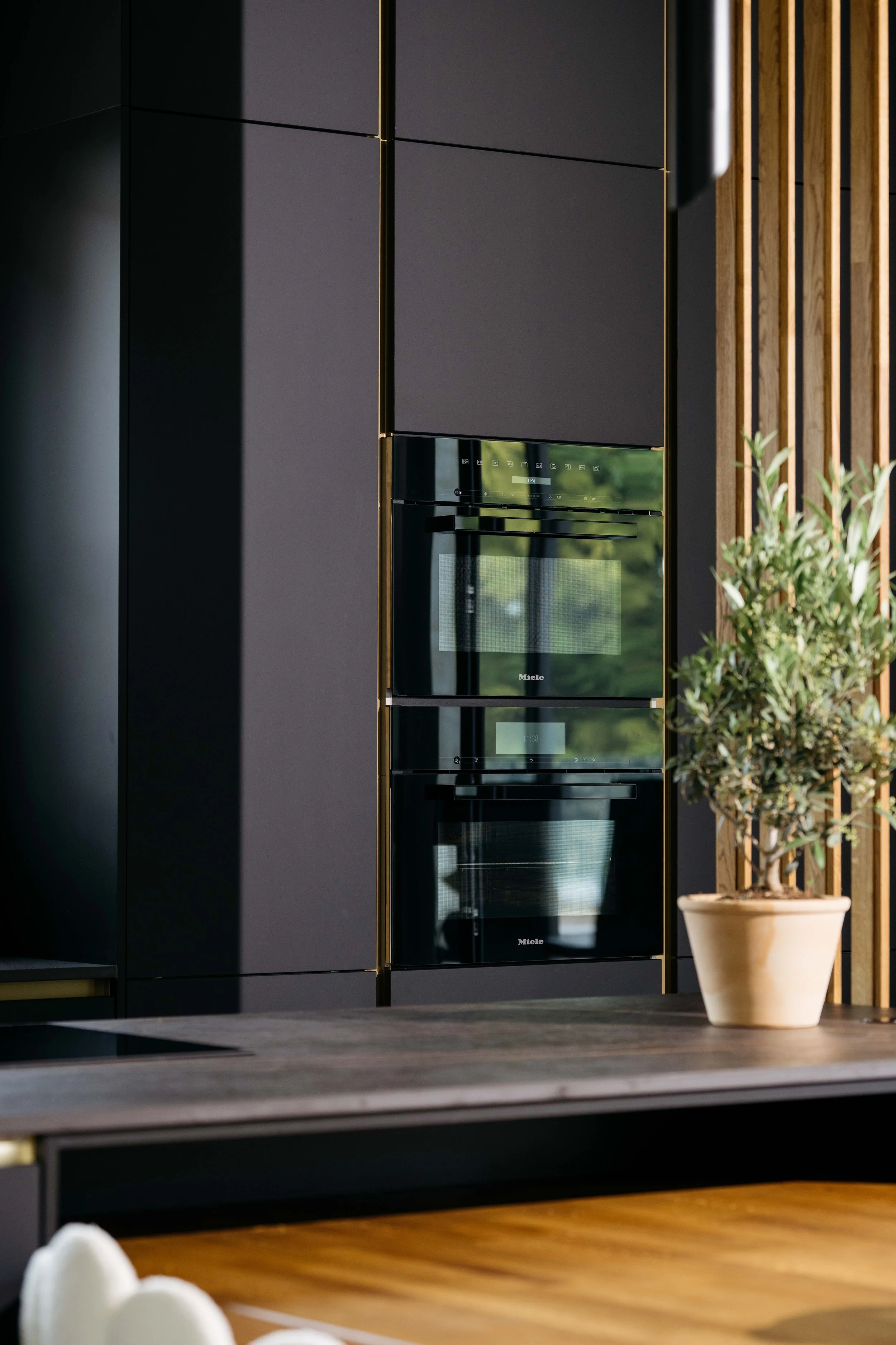 Modern inombyggnad med svarta skåp och inbyggda ugnar, samt en växt i kruka på ett träbord.