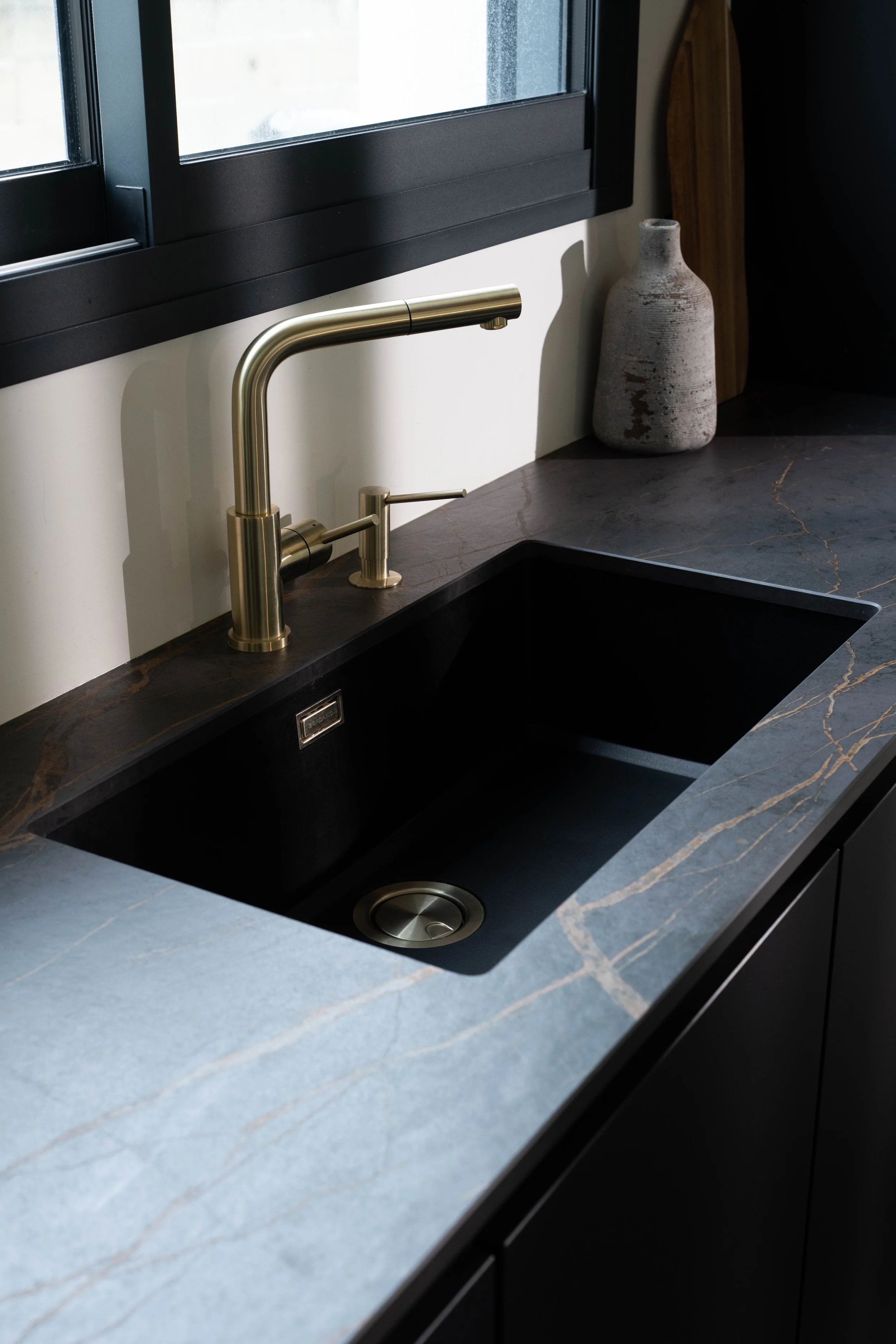 Kökssvask med svart insats och guldfärgad kran på en mörk stenbänk, vid ett fönster med svarta ramar. En dekorativ vas står på bänken.