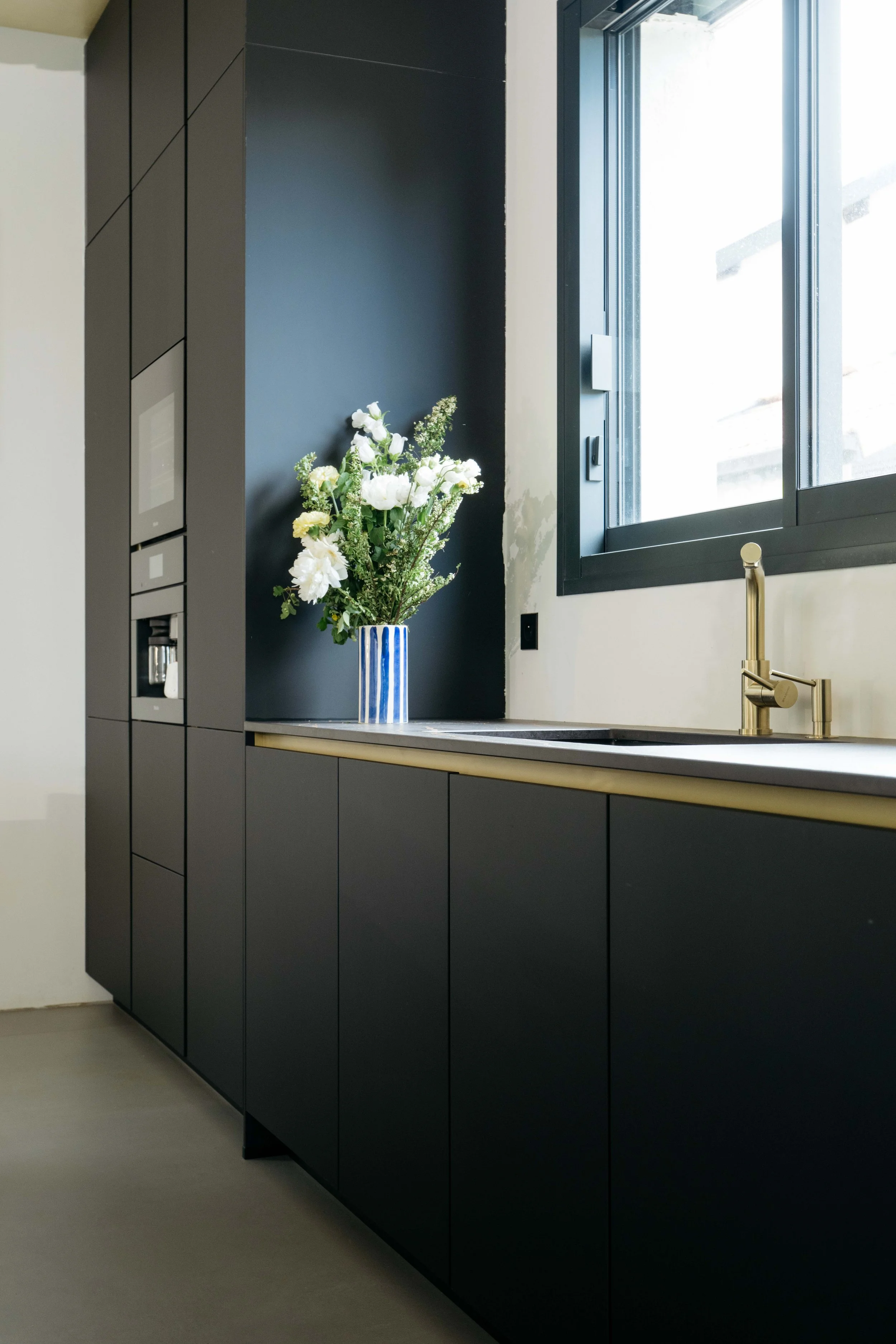 kök med svarta skåp, ett fönster, en vas med vita blommor och en guldkran.