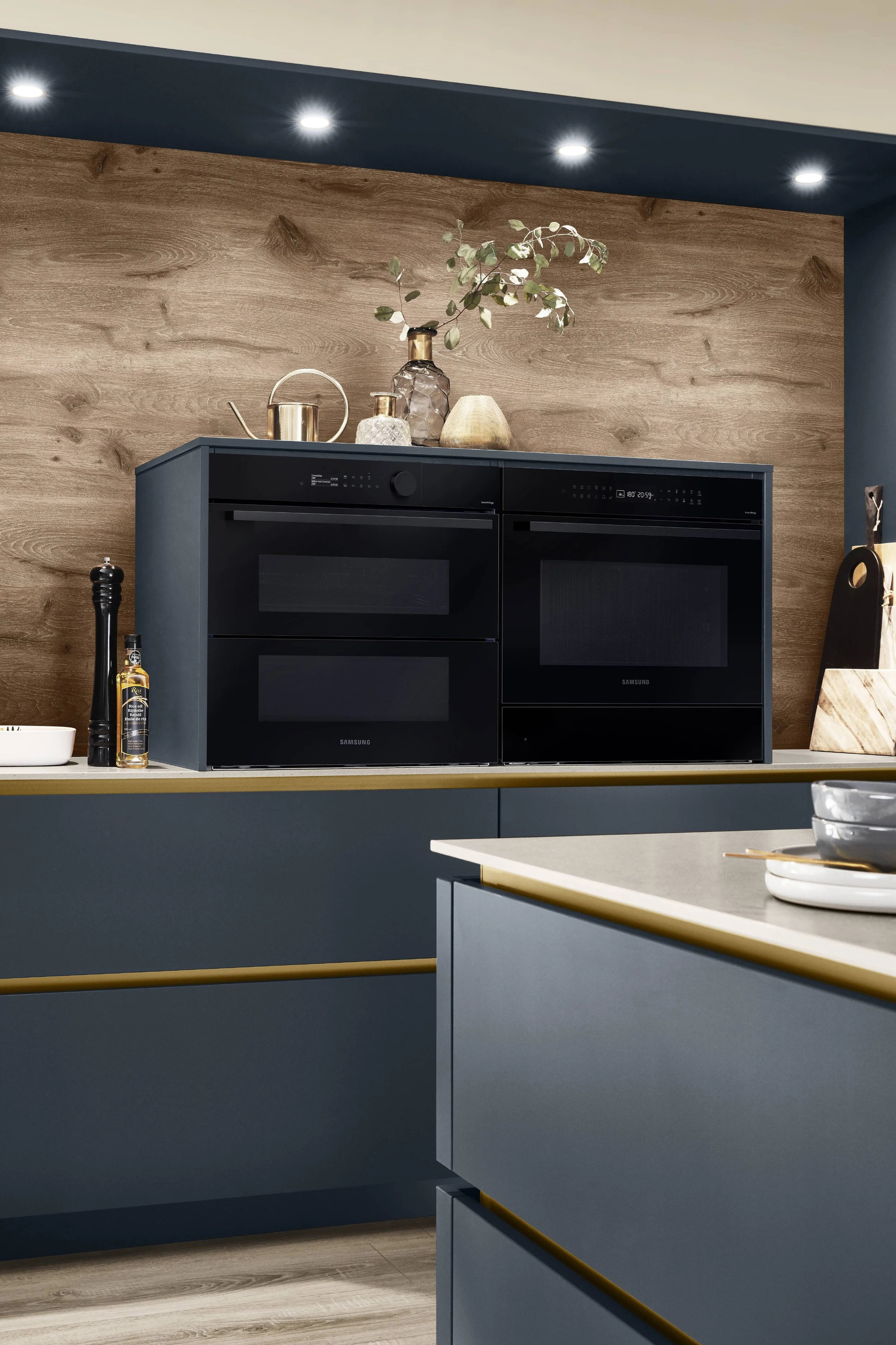 Köksö med blå skåp, tre inbyggda ugnar, dekorativa vaser och skålar, träpanelvägg, lampa i taket.