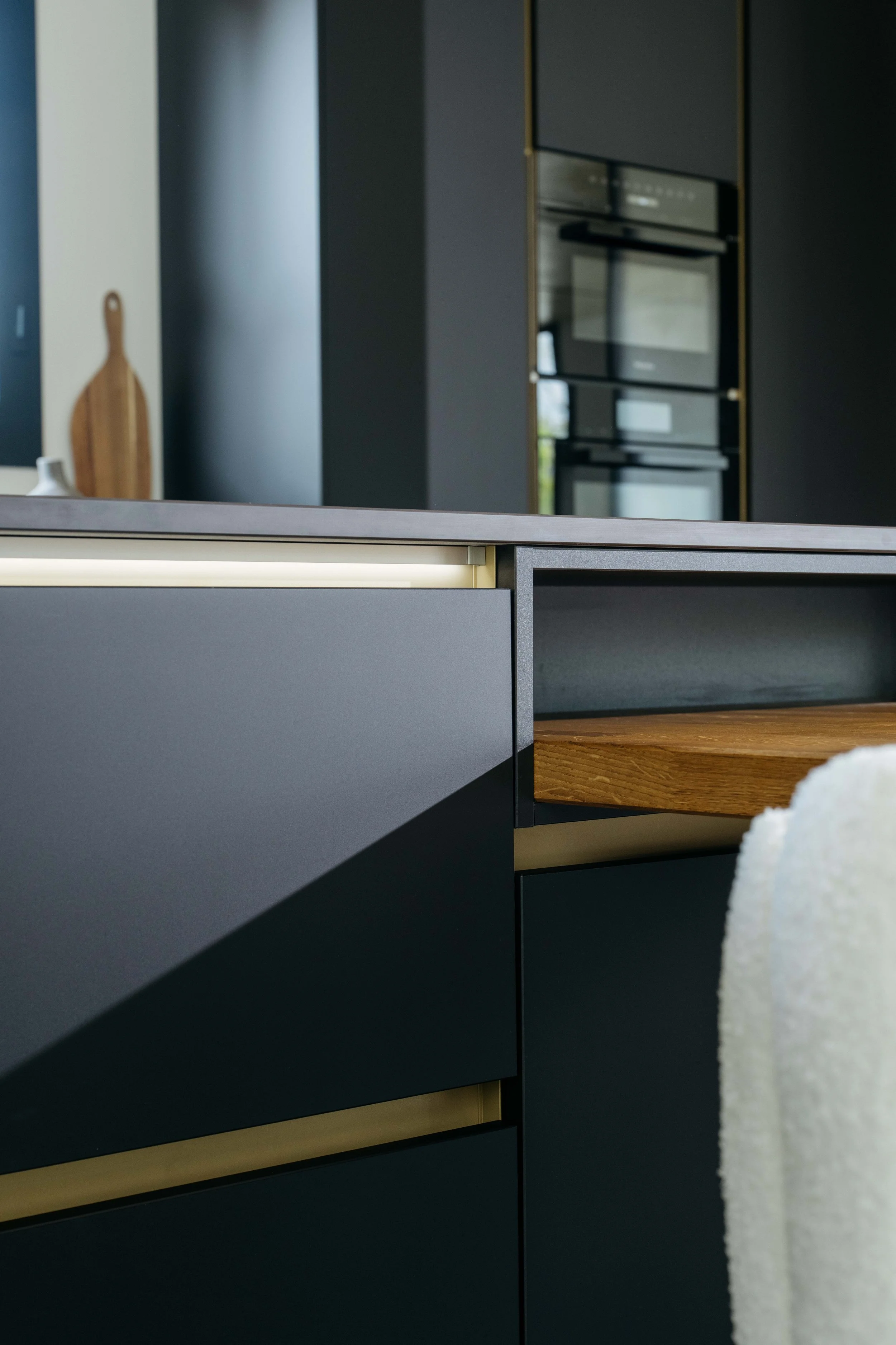 Detalj av en modern köksskåp med mörka, släta luckor och en trädetalj i ett välplanerat kök. I bakgrunden syns en mörk vägg och inbyggda ugnar.
