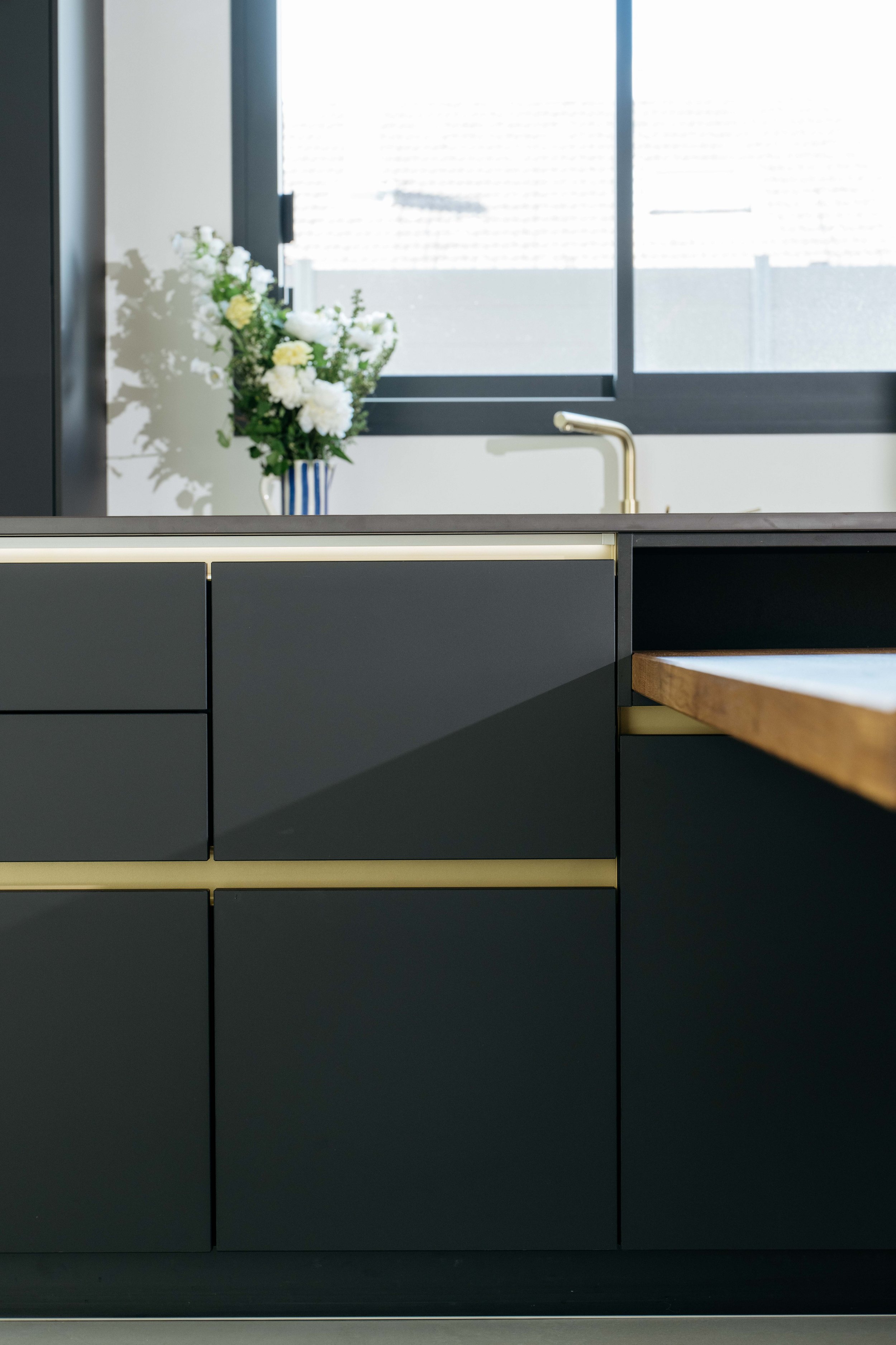 Moderna köksskåp i mörkgrått med gulddetaljer och ett trähantverk, placerat framför ett fönster med en blomsterarrangemang i en blåvit vas.