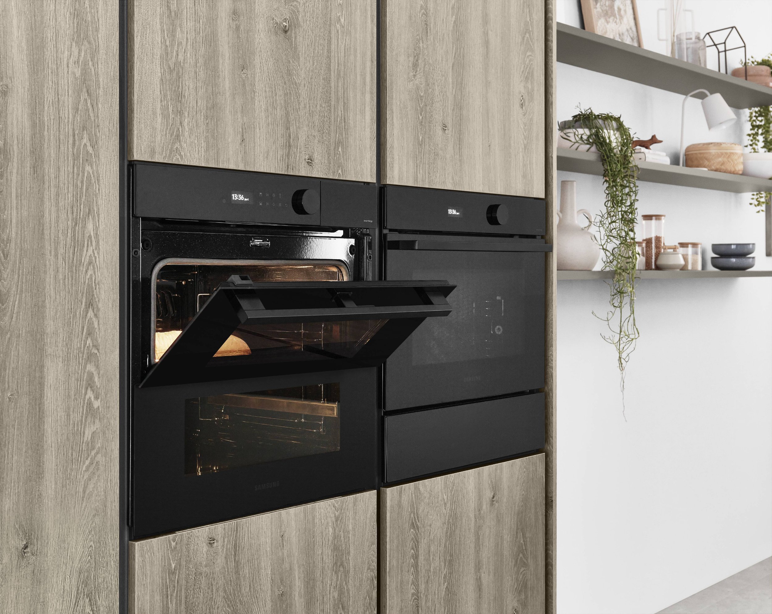 Två inbyggda ugnar i ett kök med träfasad, en har luckan öppen, och ett öppet skåp med vita och svarta skålar och växter på öppna hyllor.