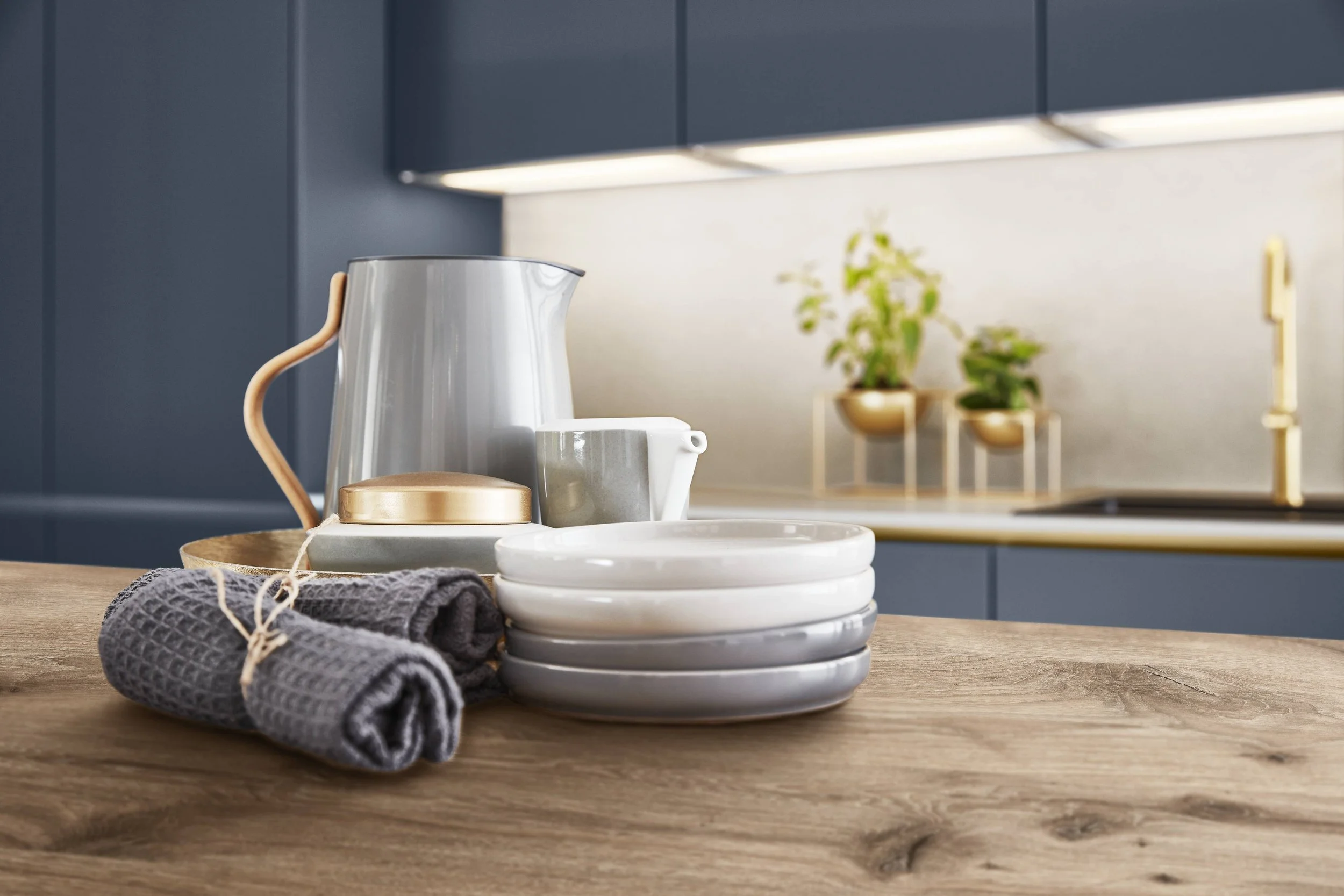 Köksbord med grå handdukar, tallrikar, kopp, kanna och förvaringsburkar, mot en blå köksskåp och växter i bakgrunden.