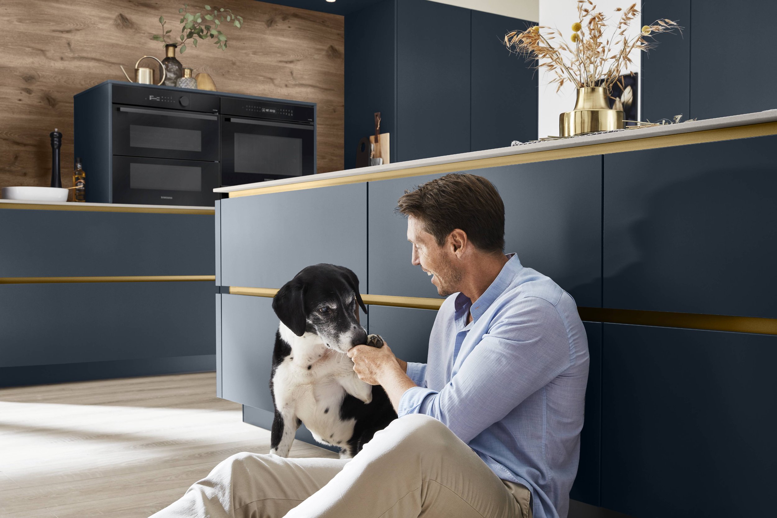 En man leker med en svart och vit hund i ett modernt kök, sittande på golvet bredvid köksbänken.