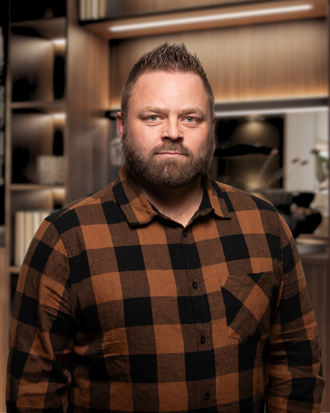 Man i rutiga brunt och svart skjorta framför kök. Det är en inomhusbild med träskåp och köksutrustning i bakgrunden.
