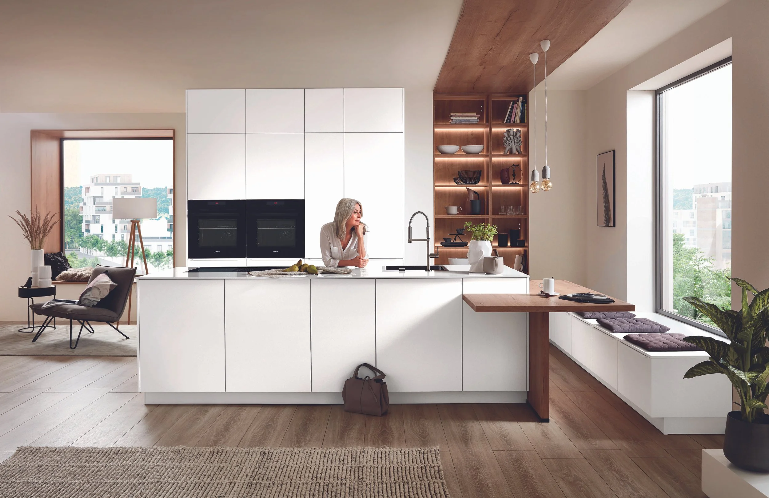 Modernt kök med vita skåp, trägolv, stor köksö med vask, sittplatser vid fönster, kvinna, stora fönster, öppen planlösning, enkelt och ljus inredd rum.
