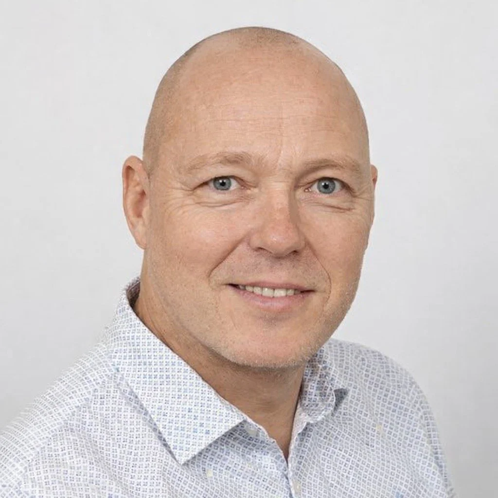 Porträtt av en man med blek hud, blå ögon, och ett kortklippt, ljust huvud, som bär en vit skjorta med ett lätt rutmönster.