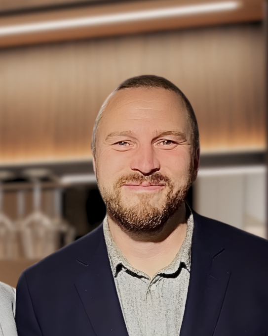 En man med skägg och kort hår, iklädd kavaj och skjorta, står i ett rum med träväggar.