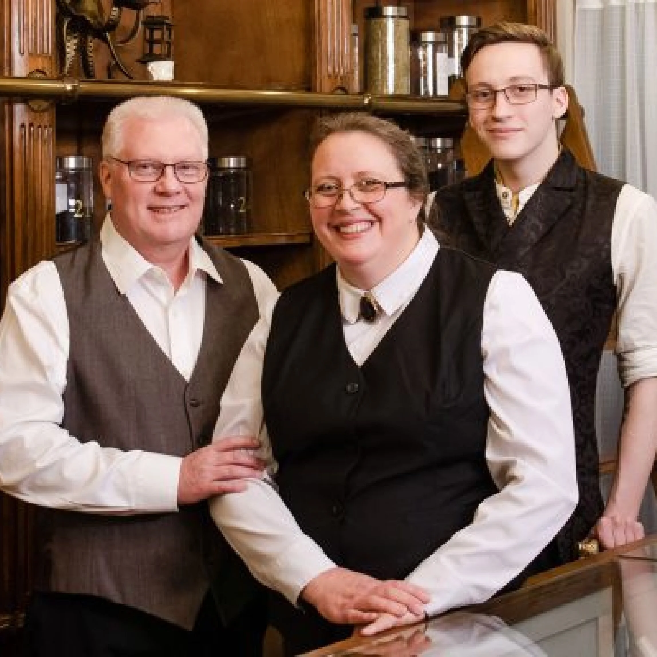 Three people dressed in vintage-style clothing standing together in a cozy, wood-paneled room. The two men and one woman are smiling and looking at the camera.