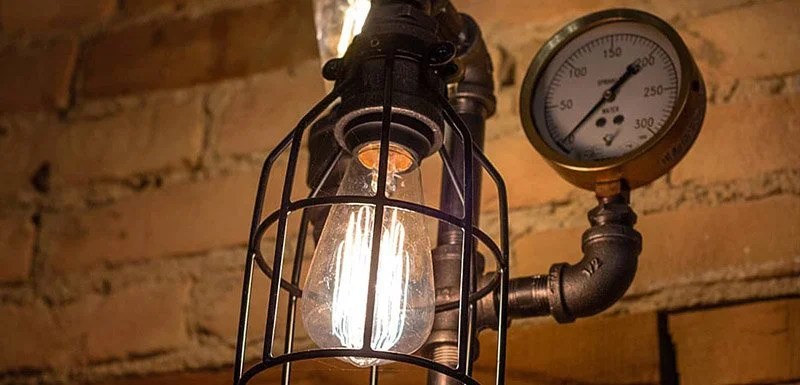 Industrial-style light bulb enclosed in a wire cage with a pressure gauge attached to the pipe next to it against a brick wall background.