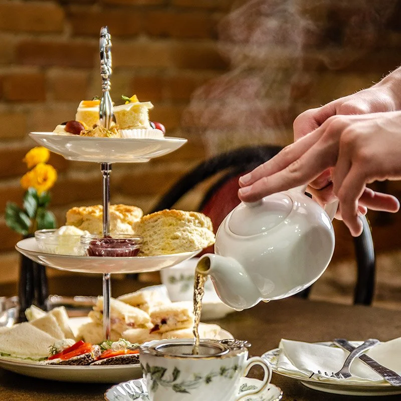 A person pouring tea into a cup with a three-tiered serving tray of assorted desserts and sandwiches in the background.