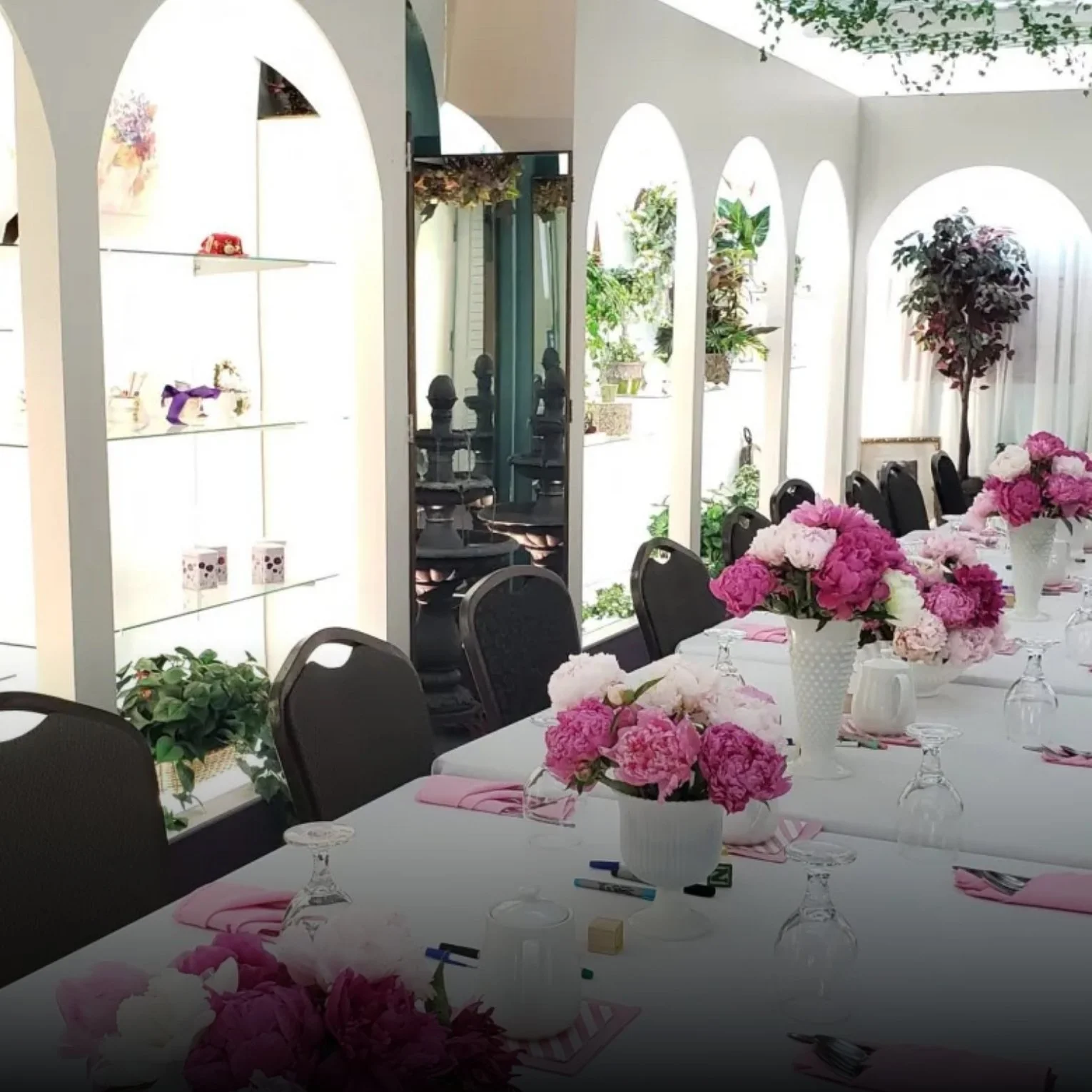 A decorated dining table set with pink and white flower arrangements in vases, pink napkins, and glasses, inside a bright room with arched windows and planters with greenery.