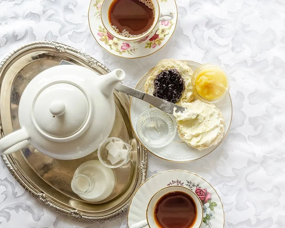 Tea set with teapot, cups of tea, and a plate of scones with jam, clotted cream, and butter on a table with a white tablecloth.