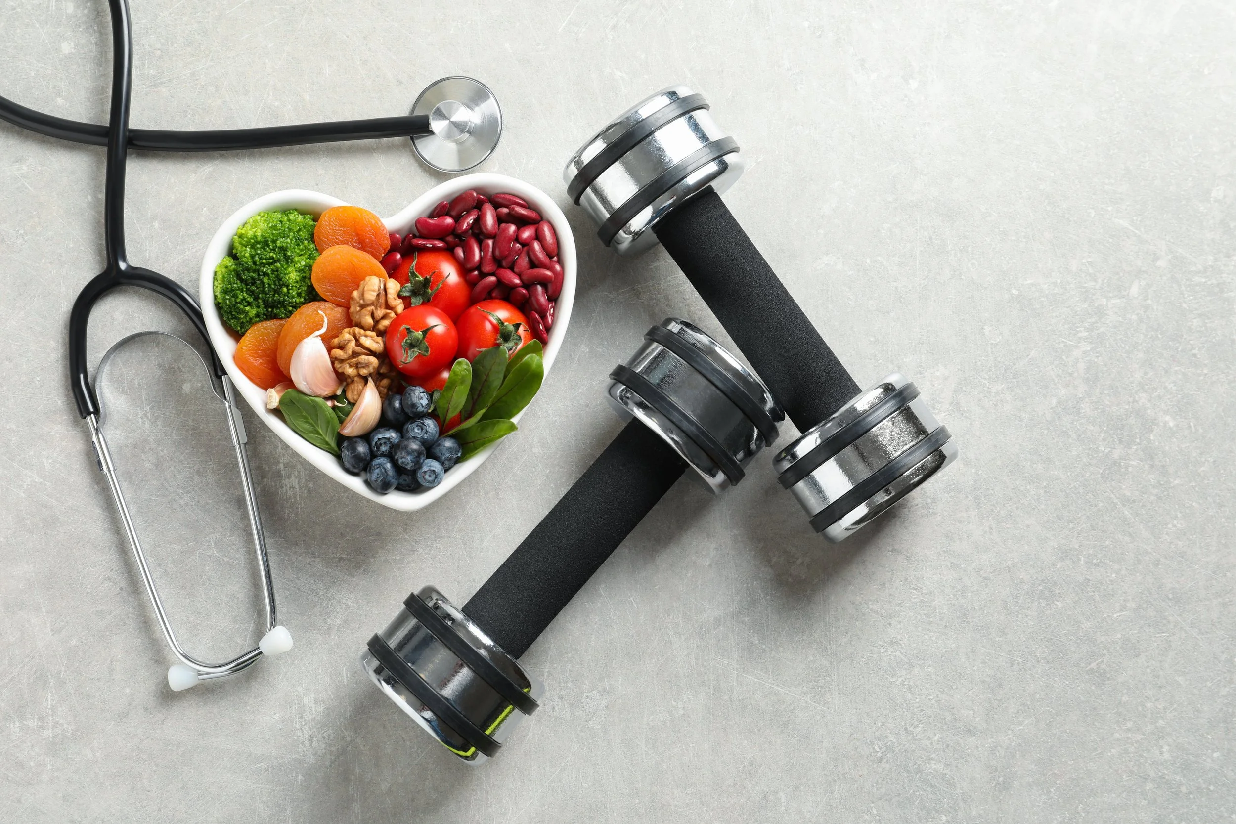 A heart-shaped bowl filled with fresh vegetables, berries, and nuts next to an adjustable dumbbell and a stethoscope on a light grey surface.