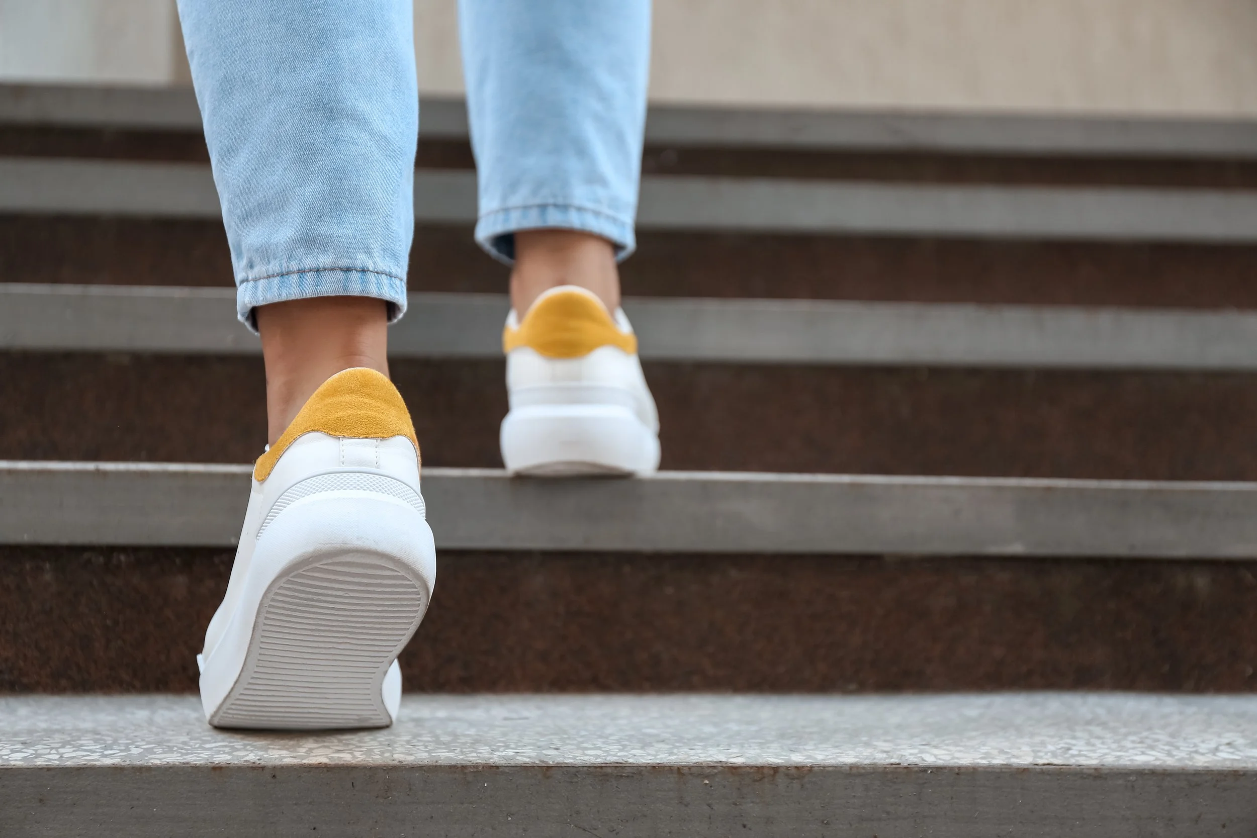 Person wearing white sneakers with yellow accents and light blue jeans climbing outdoor stairs.