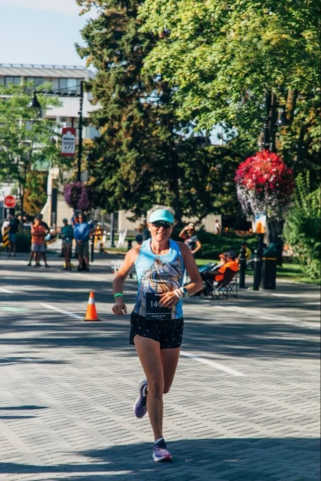 Woman running in a marathon event, smiling, wearing sunglasses, a tank top with a design, and shorts, on a city street with trees, hanging flower baskets, and spectators in the background.