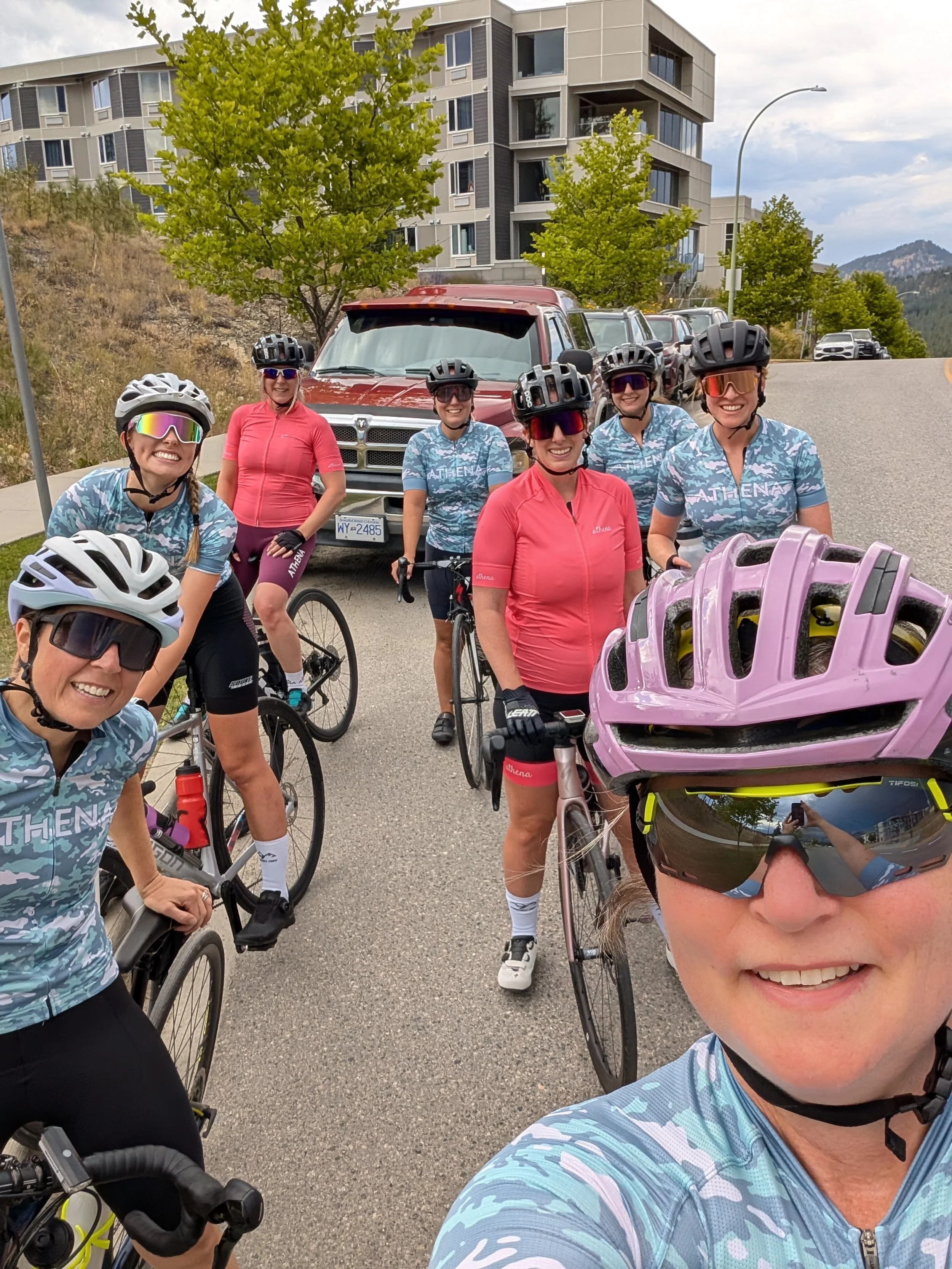 A group of seven women cyclists wearing helmets and sunglasses, posing with their bikes outdoors on a road with parked cars behind them. They are dressed in matching blue and pink cycling jerseys, and some are smiling at the camera. In the background, there are trees, a modern building, and mountain scenery.