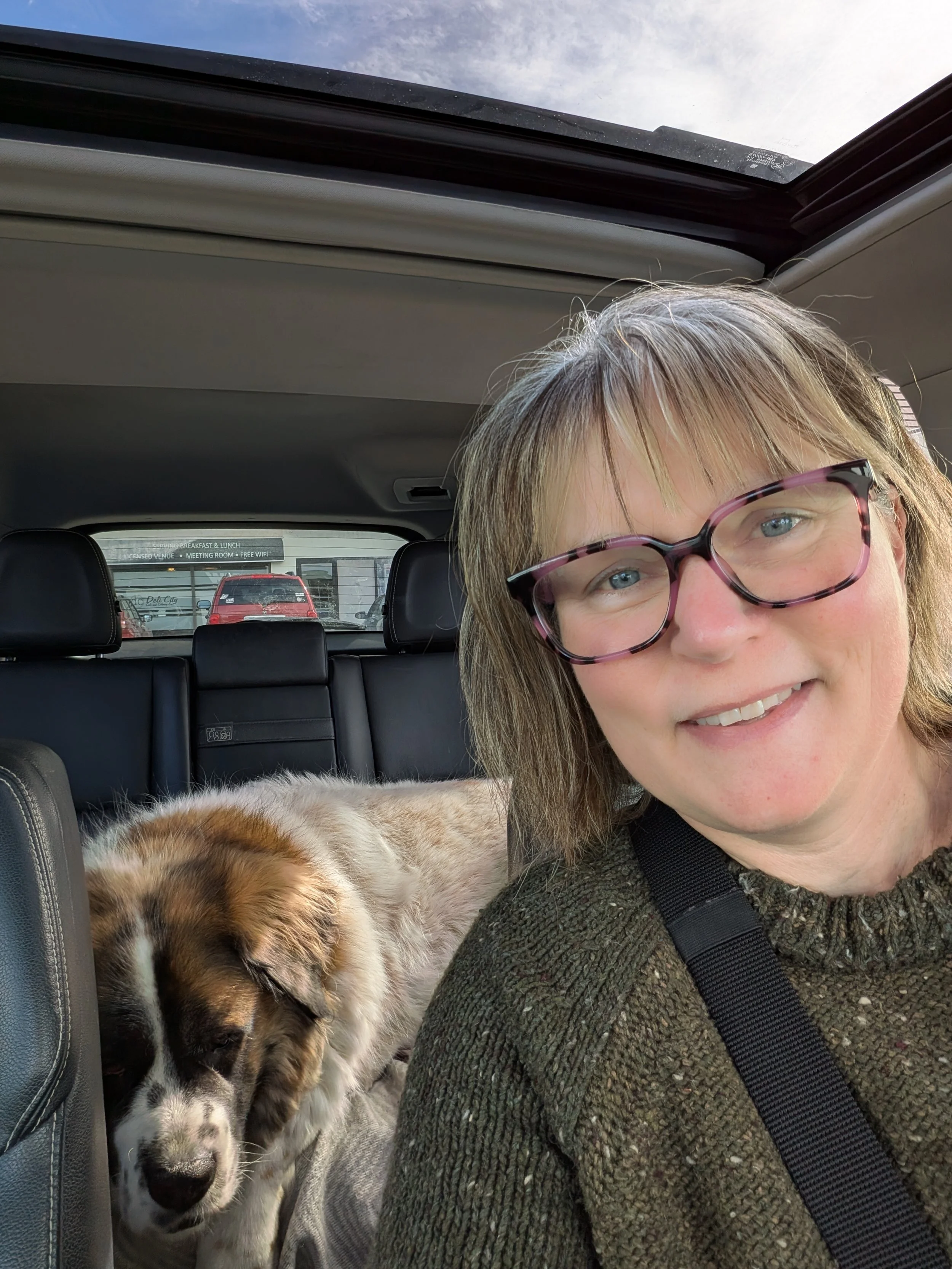 A woman with glasses smiling in the front seat of a car, with a sleeping dog lying behind her in the back seat.