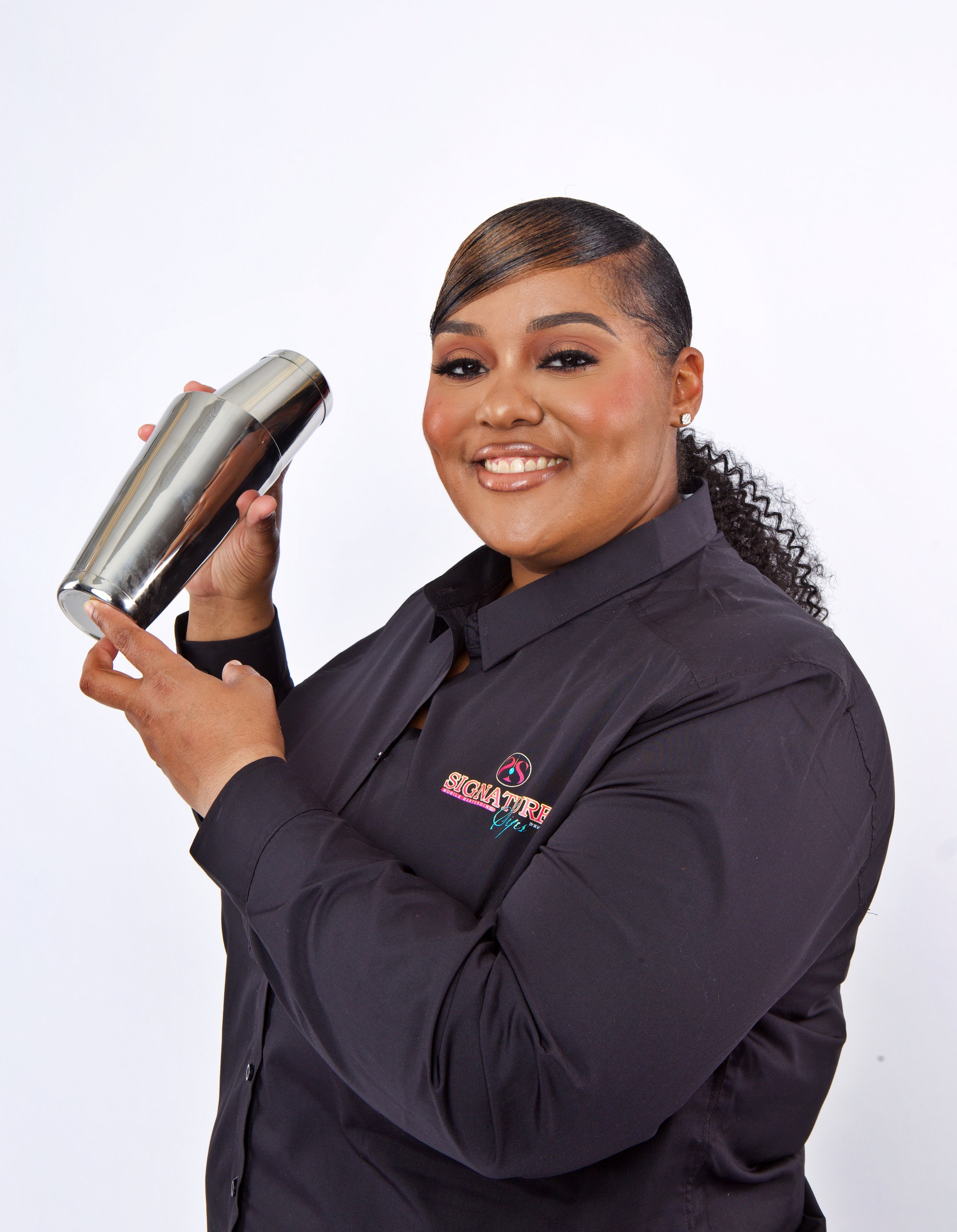 A woman with dark hair styled in a ponytail, smiling and holding a stainless steel cocktail shaker, wearing a black shirt with a logo that says 'Signature Girls'.