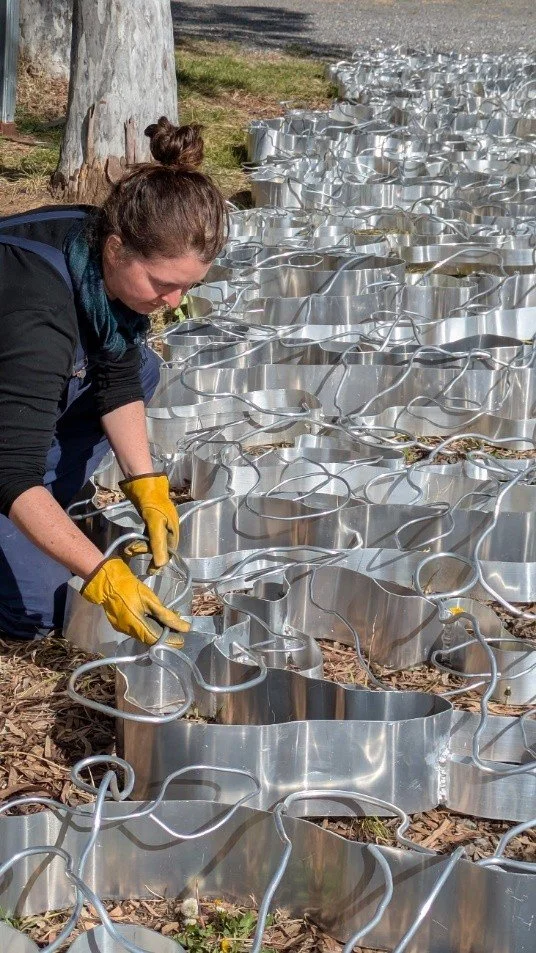 A person wearing yellow gloves adjusts reflective metal shapes laid on the ground in a natural setting, conveying a sense of focus and creativity.