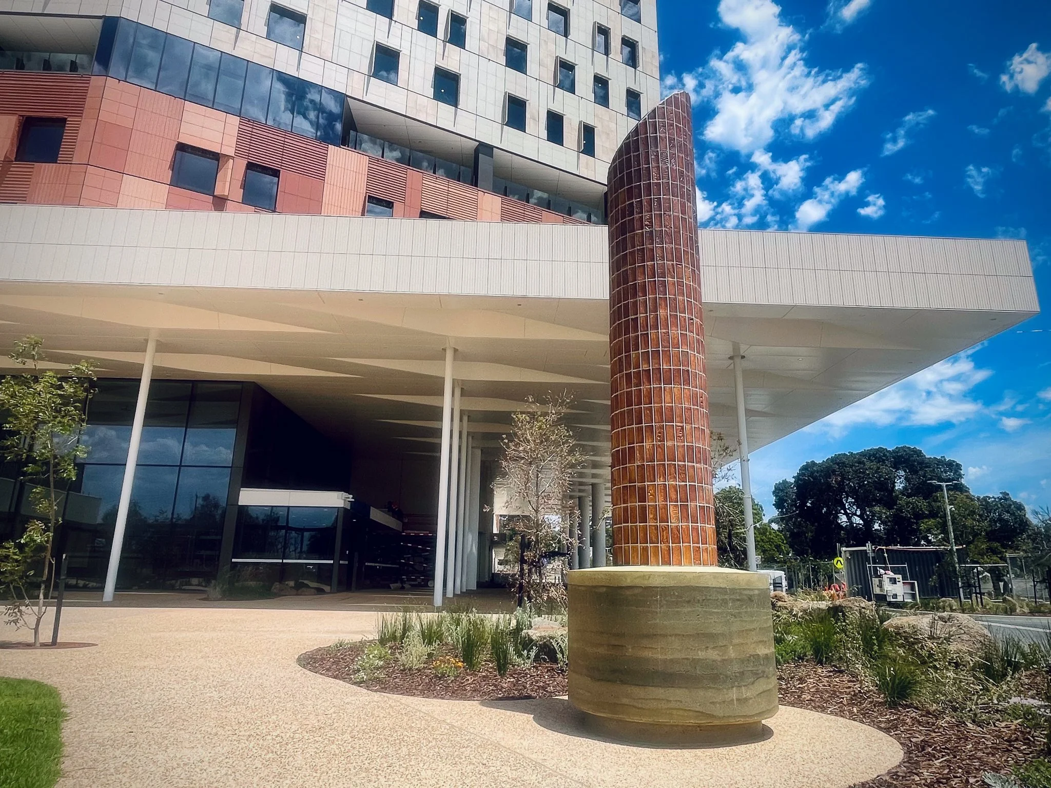 A tall, curved brick sculpture stands in front of a modern building with geometric patterns and large windows. It's set on a landscaped plaza under a blue sky.