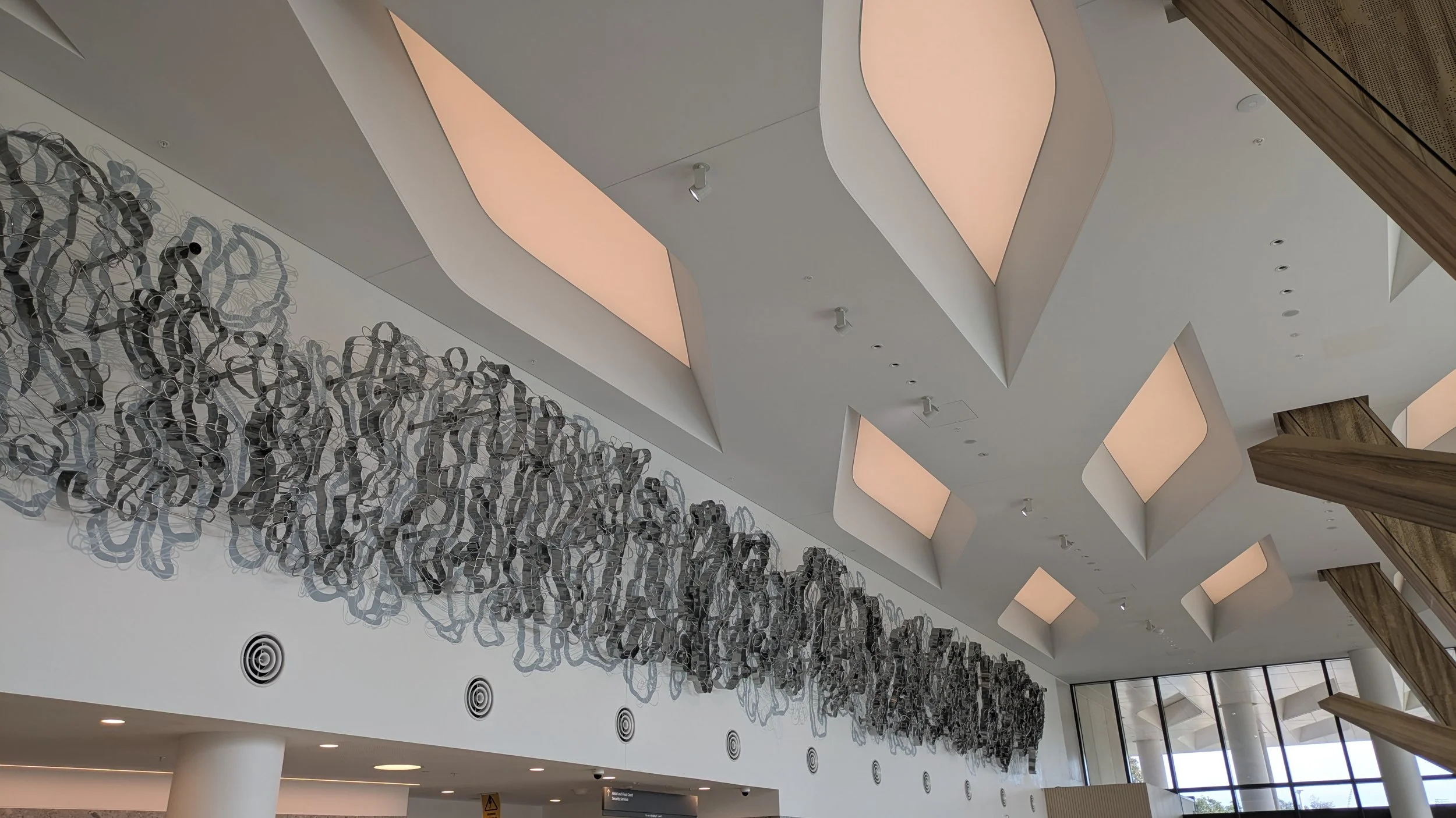 A large, intricate metal sculpture with intertwined, swirling forms hangs on a wall in a modern space. The ceiling features abstract, geometric skylights.