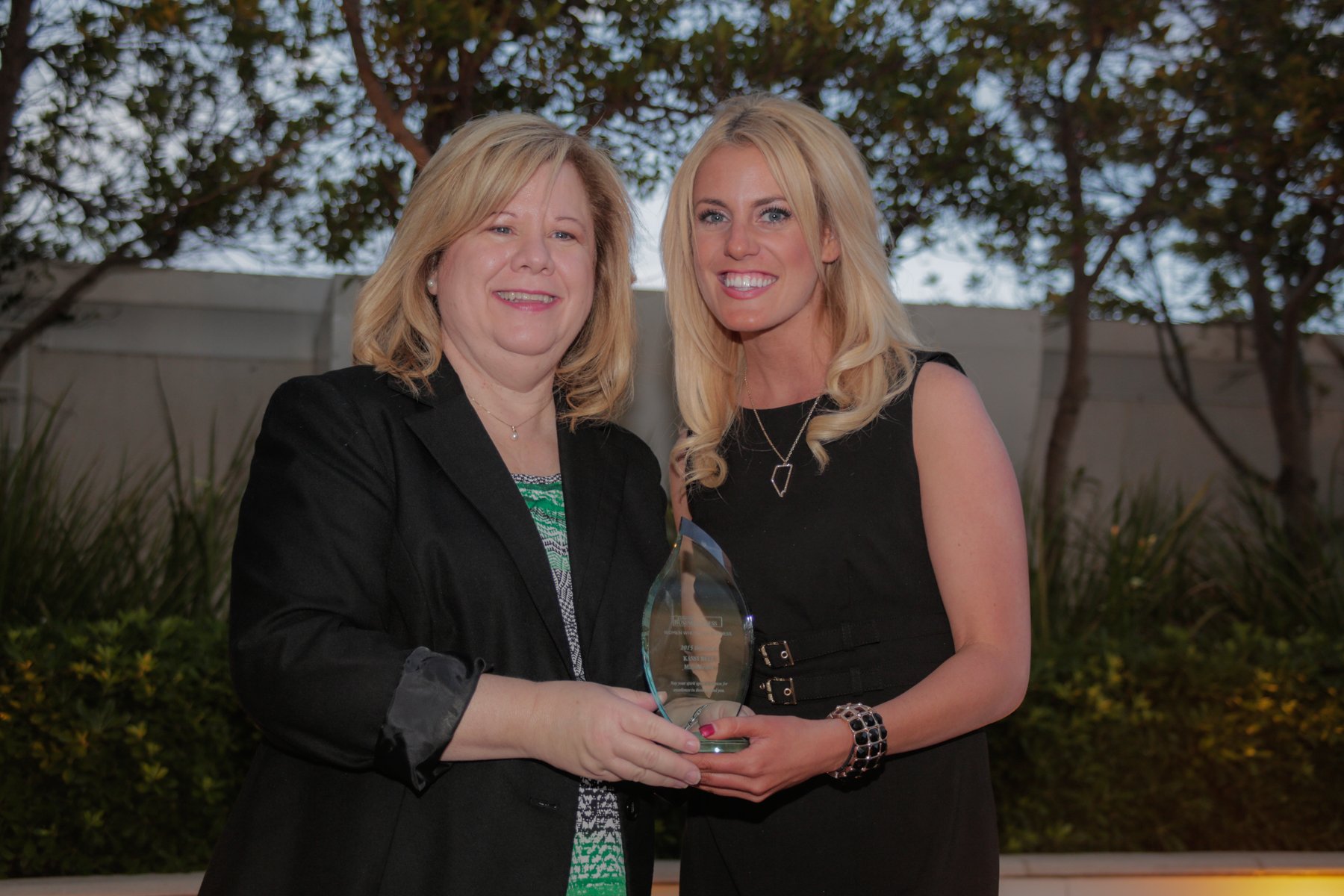 Two women standing outdoors in front of trees, holding a glass award and smiling at the camera.