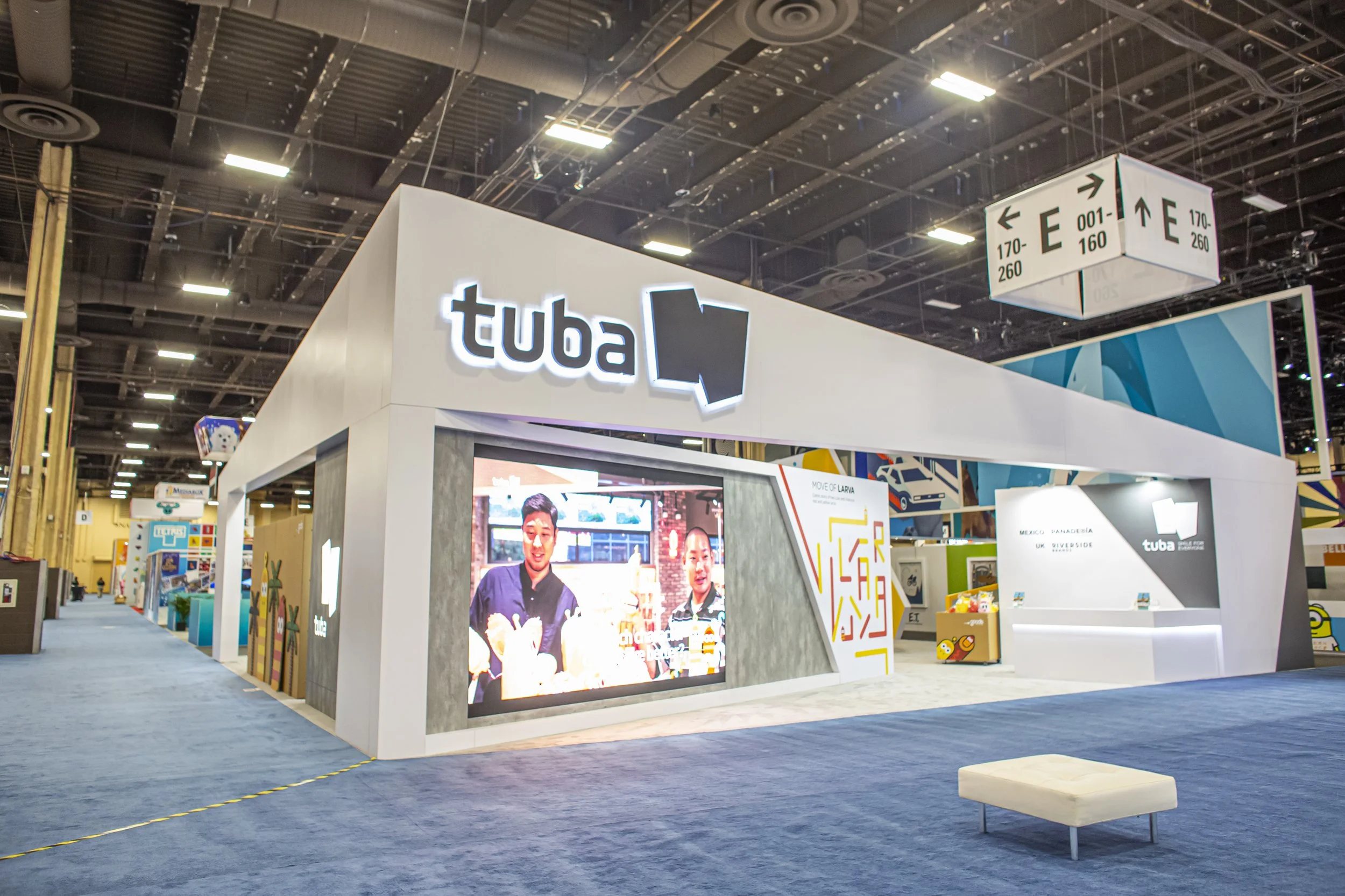Trade show booth for Tuba, featuring a large digital screen showing two people, with the Tuba logo and signage in a spacious exhibition hall.