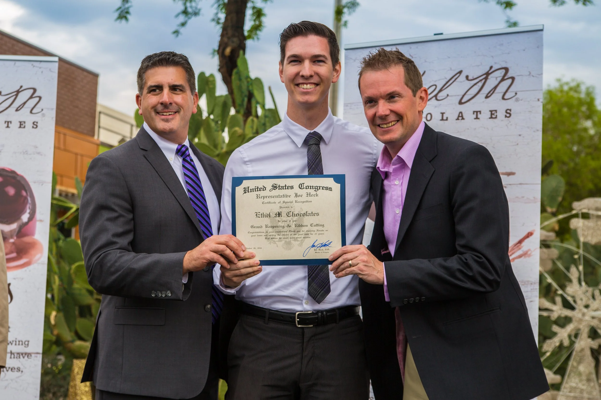 Three men in suits holding a certificate, celebrating an award presentation outdoors. Behind them are banners and large cactus plants.
